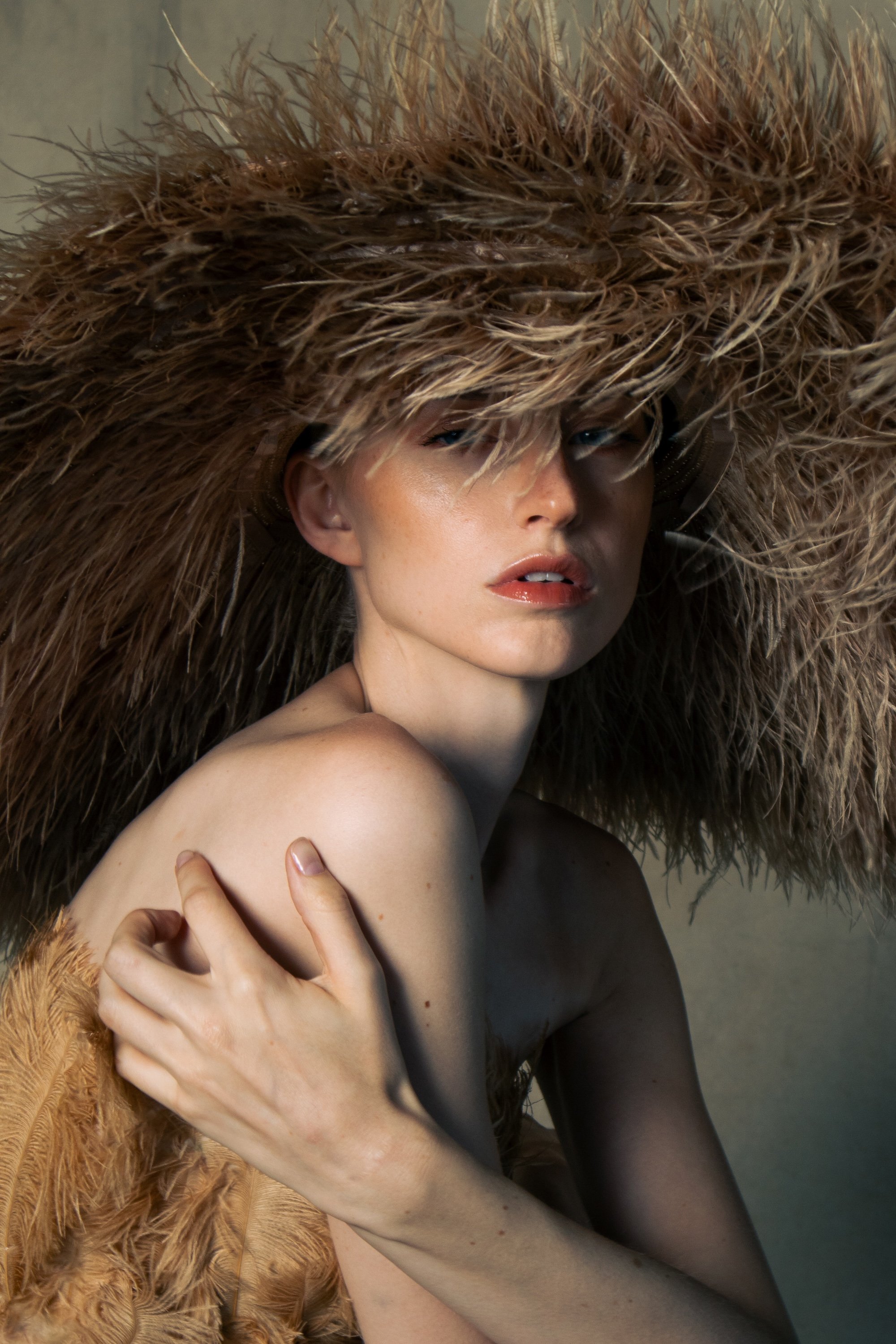 A woman with fair skin wearing a large, fluffy feathered hat covering her head and shoulders, looking at the camera with a neutral expression, bare shoulders, and holding her arm across her chest.