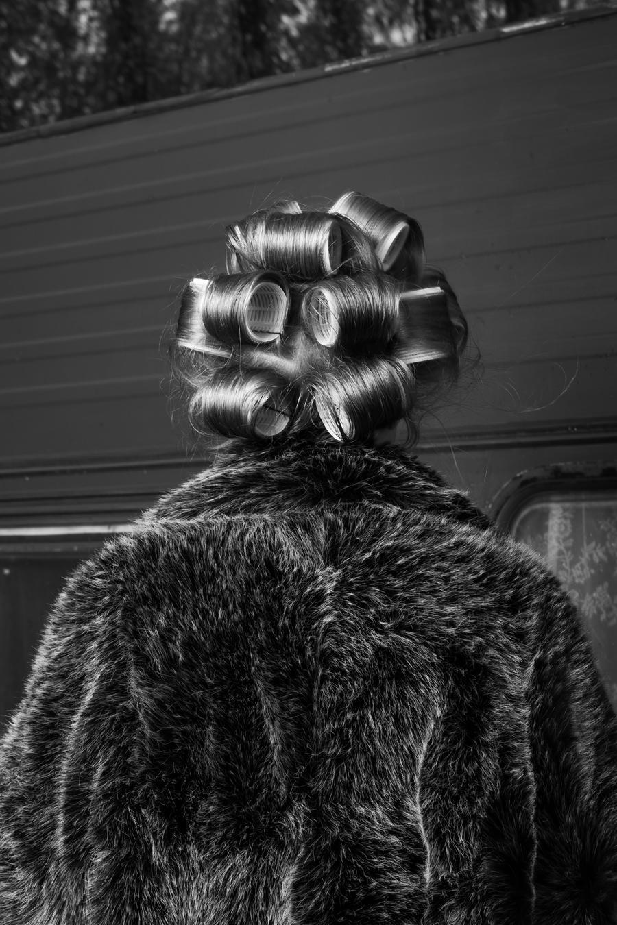 Back of a person with hair in large curlers, wearing a fur coat, outdoors near a building with trees in the background. In black and white.