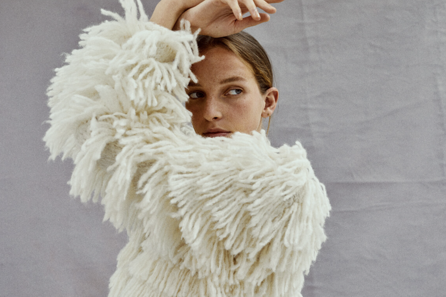 A woman with light brown hair and freckles, wearing a fluffy, cream-colored textured coat, poses with her arm raised above her head, against a light gray background.