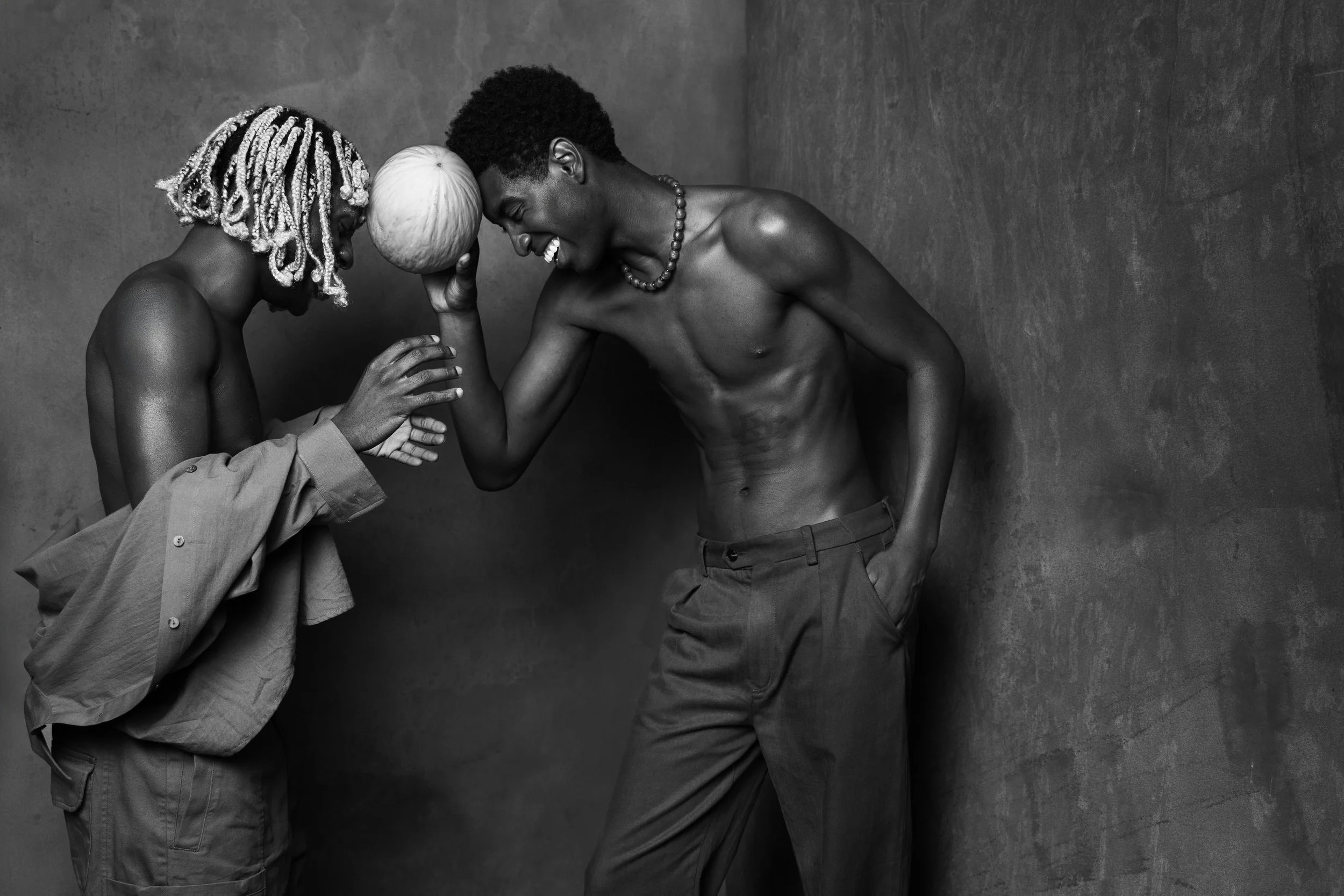Two men, one with dreadlocks and the other with short curly hair, smiling and touching foreheads. The man with dreadlocks is holding a round object near his head and is wearing a loose shirt, while the other man is shirtless, wearing a beaded necklace, and has one hand in his pocket.