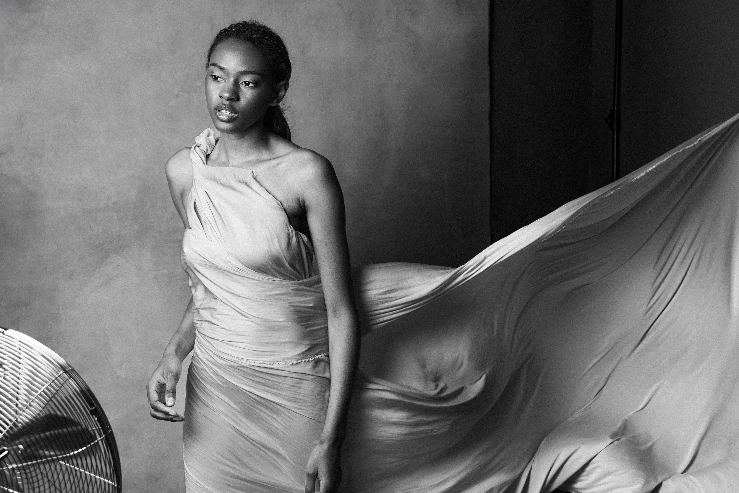 Black and white photo of a woman wearing a flowing, strapless dress, standing in a studio with a neutral background, with a fan visible in the foreground.