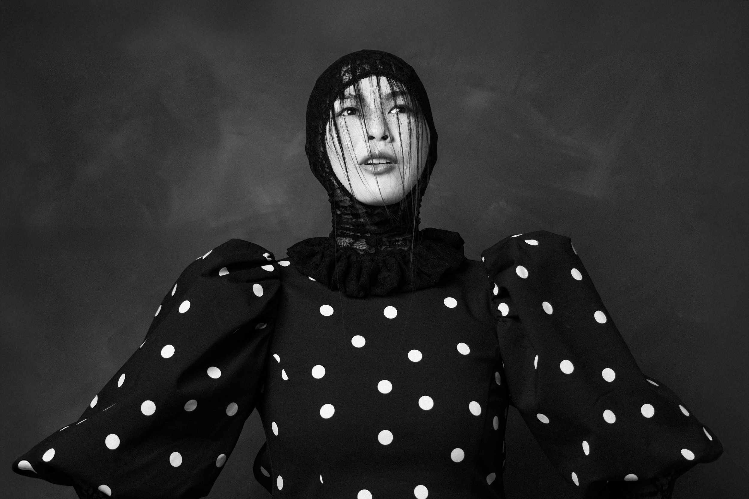 A woman wearing a polka-dot dress with puffed sleeves and a lace hood, her face partially covered by a lace veil, looking upward against a cloudy sky.