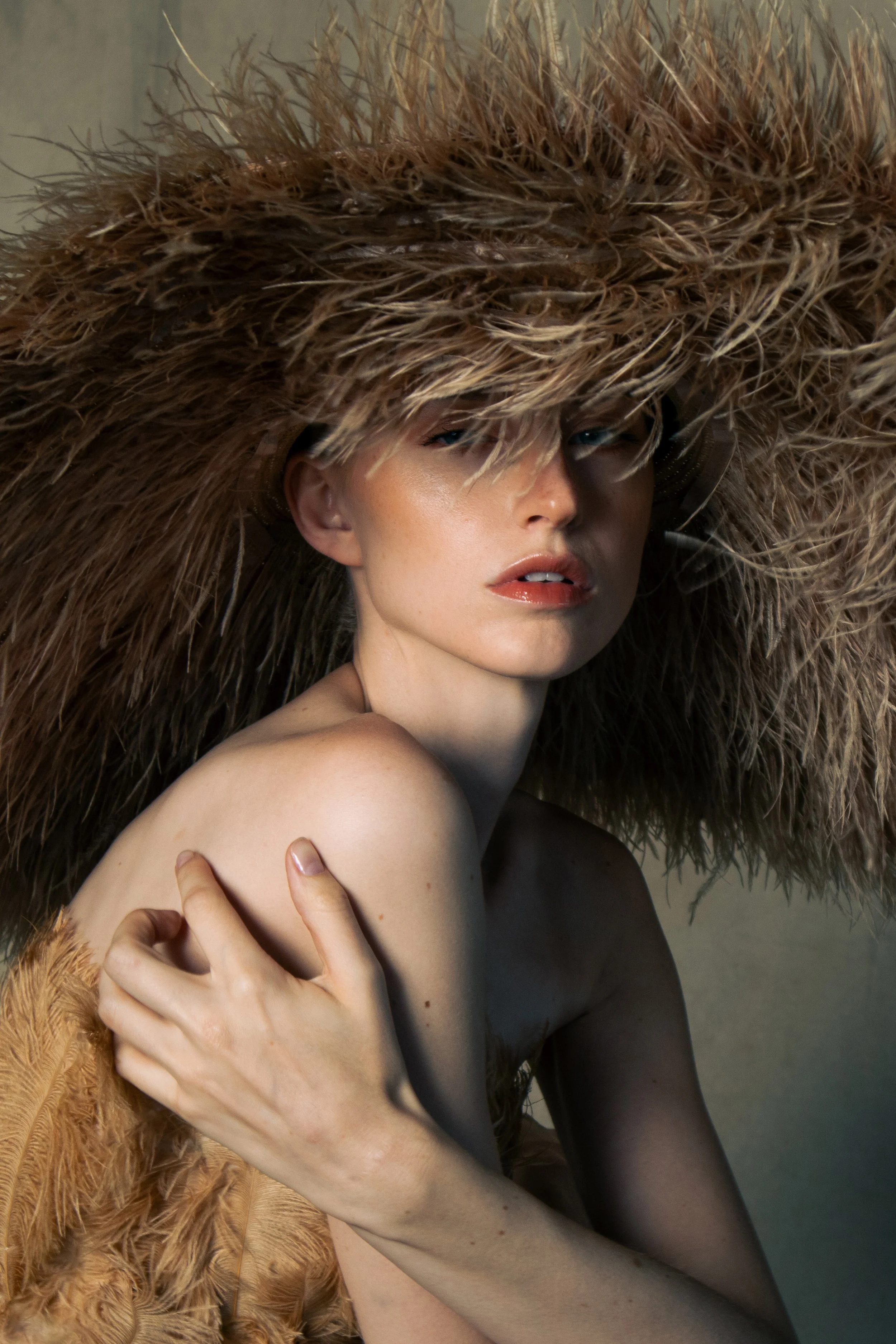 A woman wearing a large, fluffy brown hat made of feathers and fur, covering her head and partly obscuring her face, with her arm crossed over her chest.