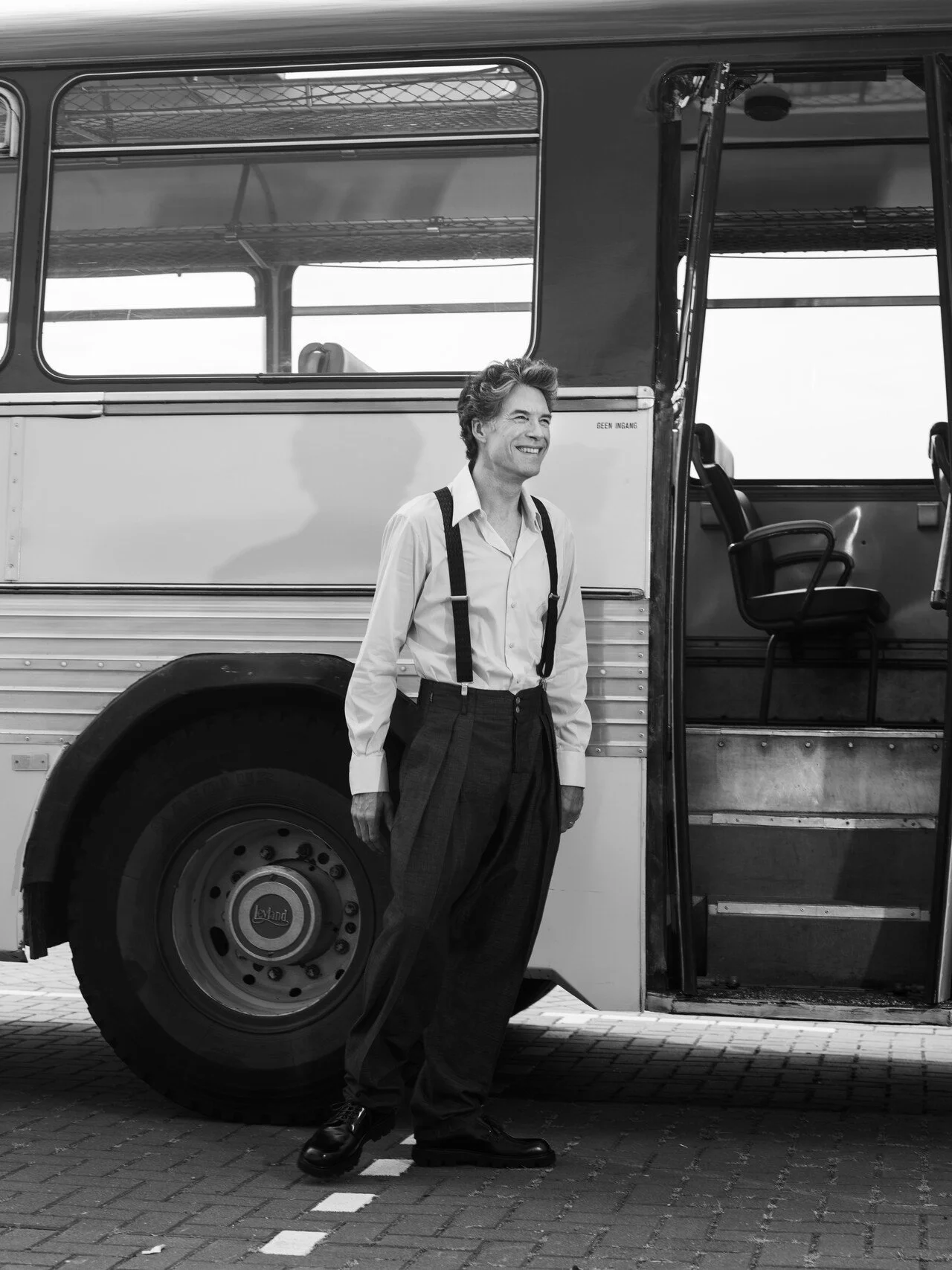 A man standing next to a vintage bus, smiling, dressed in a white shirt, suspenders, and dark pants, with the bus's open door to his right.