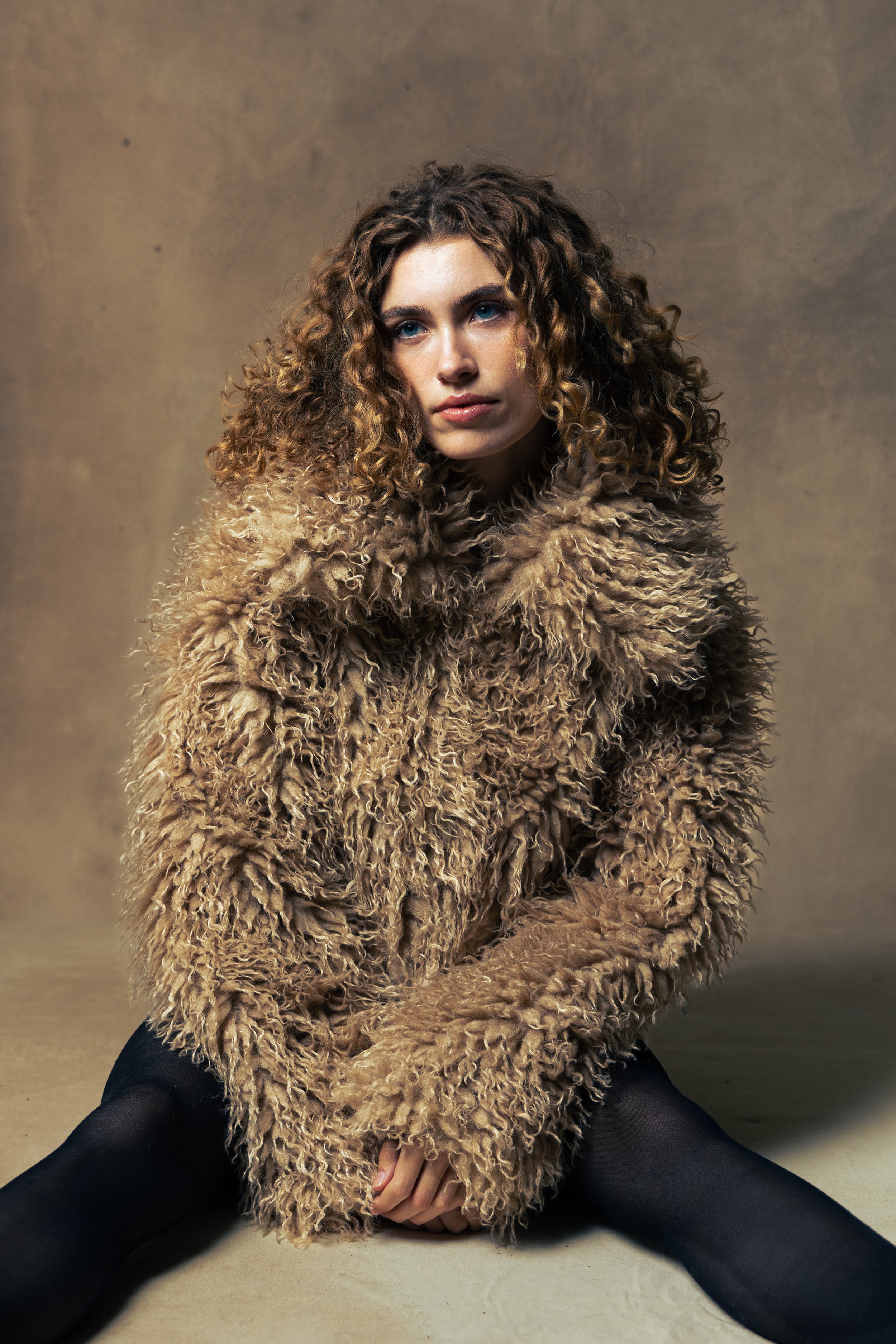 Young woman with curly hair wearing a large, fluffy beige coat with black leggings, sitting on the floor against a plain background.