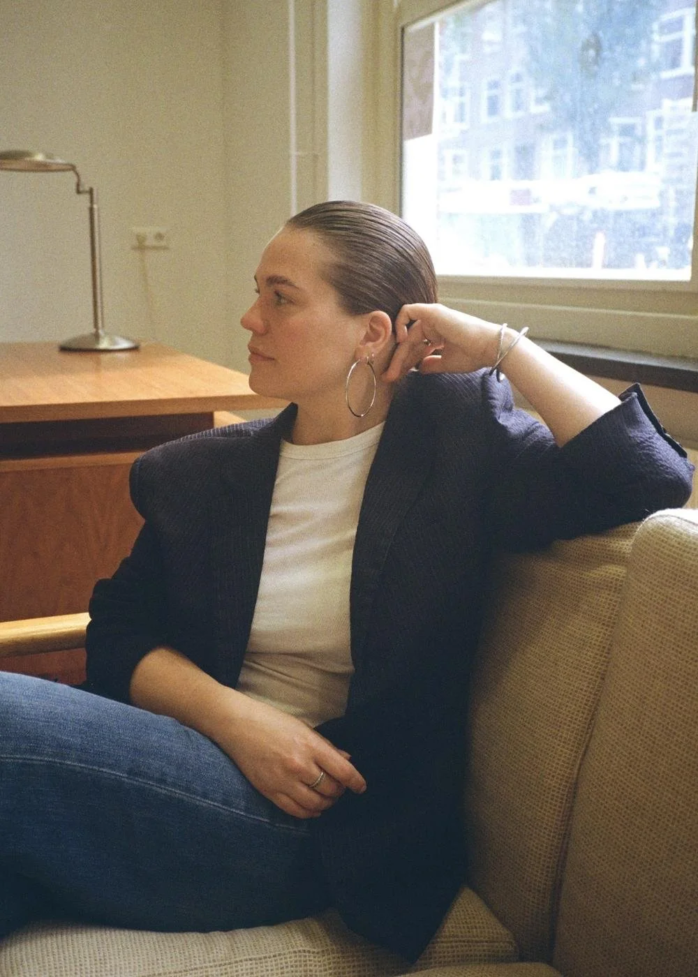 A woman sitting on a beige couch near a window, with her hair slicked back, wearing a dark blazer, white t-shirt, hoop earrings, and a bracelet, in a well-lit room with a wooden desk and lamp in the background.