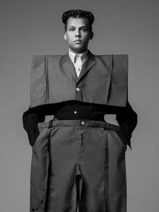 Black and white photo of a person wearing a suit with oversized, exaggerated pants and shoulders, standing with hands in pockets.