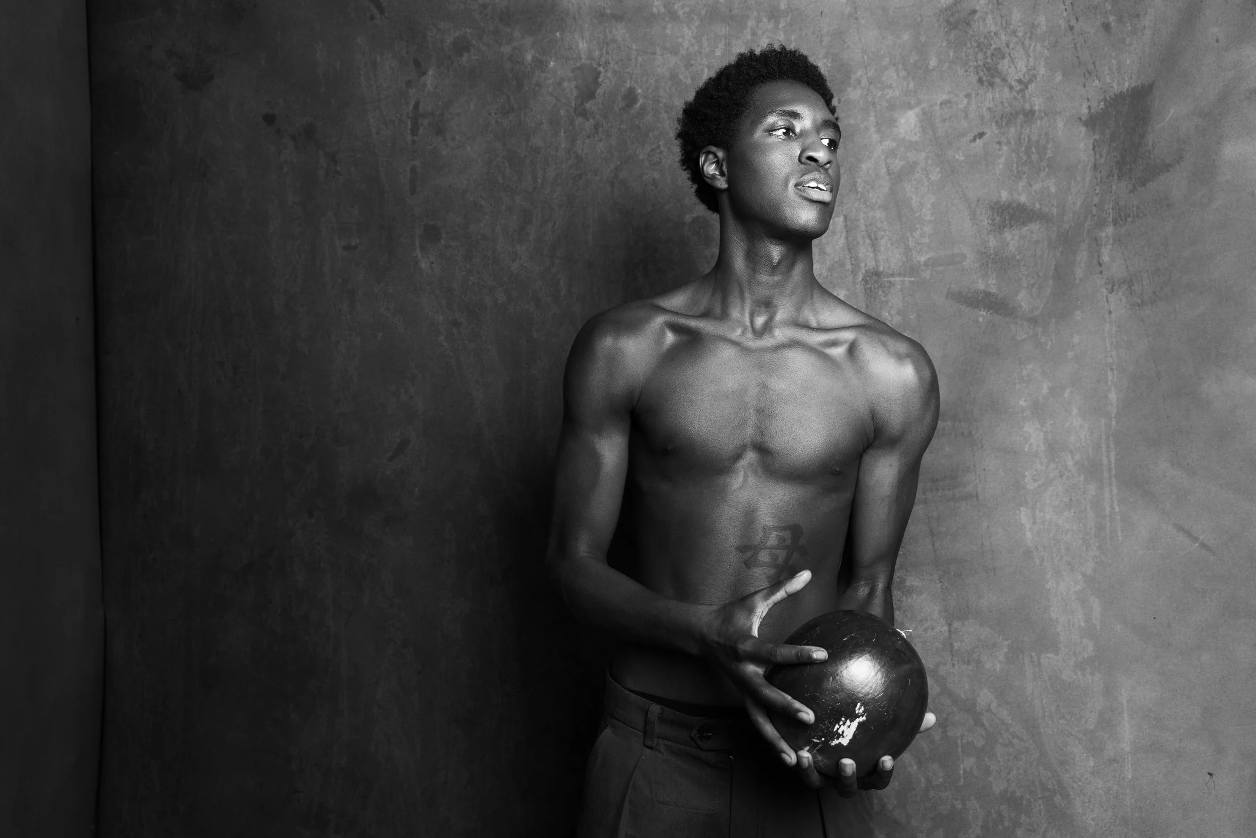 A shirtless man with short curly hair holding a black spherical object against a textured wall, looking to the side in a black and white photograph.