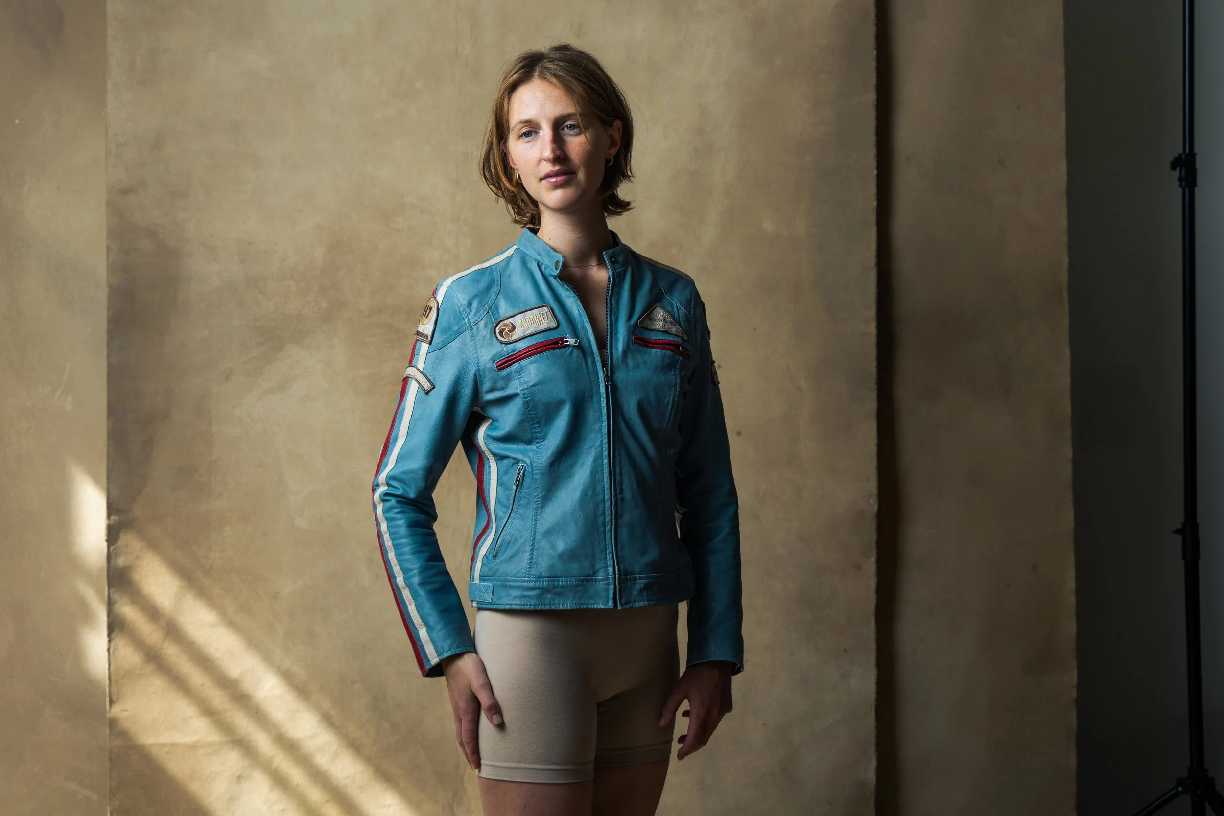 A young woman with short light brown hair wearing a vintage blue racing jacket with patches and beige shorts.