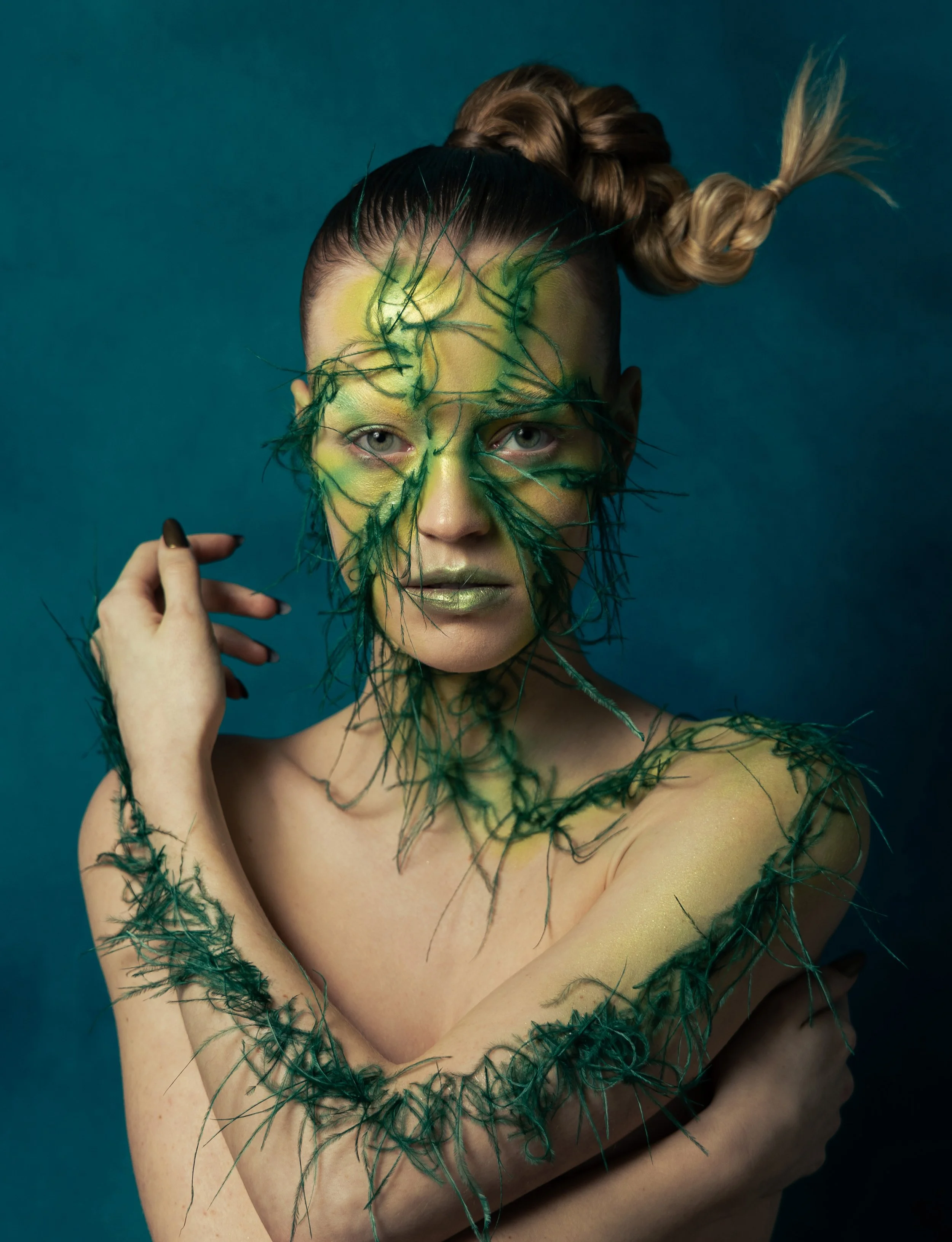A woman with eye makeup and green feather art covering her face and body, styled with an updo hairstyle, against a teal background.