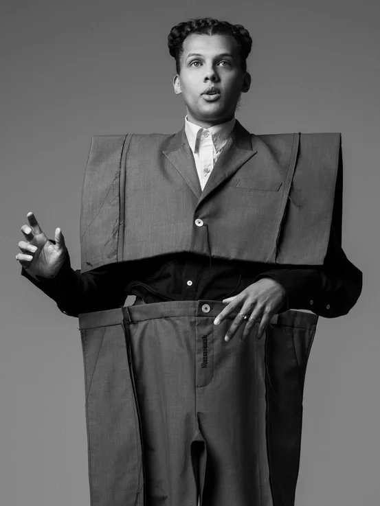 A black and white photo of a woman wearing a suit, with oversized suit pieces wrapped around her body and arms, standing against a plain background.