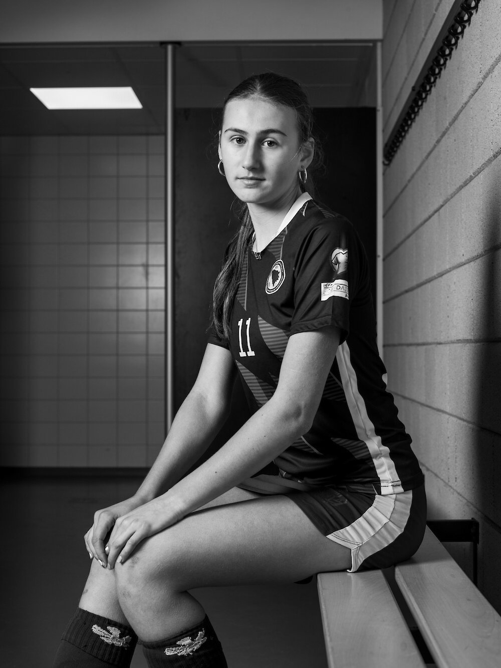Black and white photo of a young woman in a sports jersey sitting on a bench in a locker room.