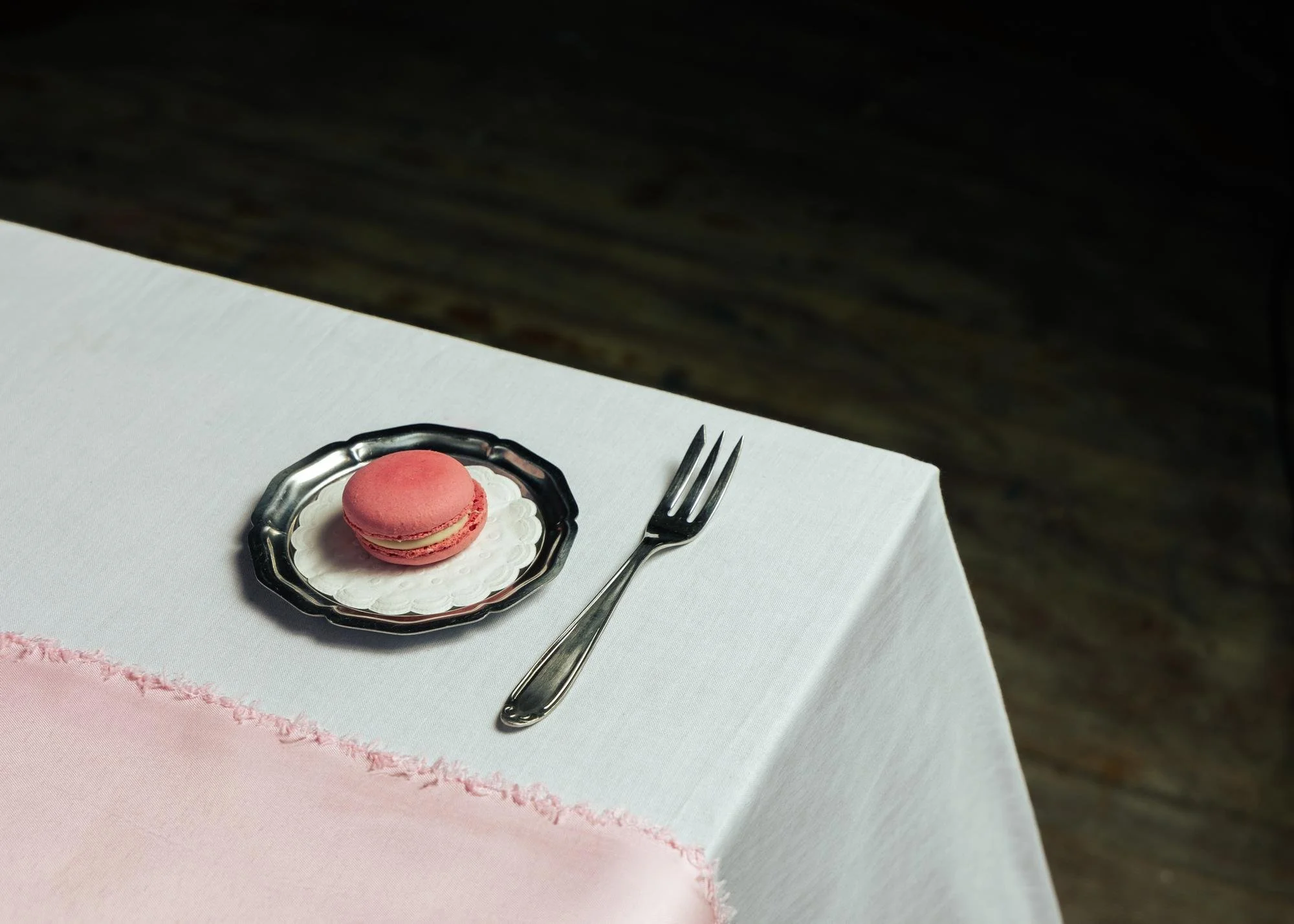 A pink macaron on a small silver plate with a paper doily, next to a silver fork on a white tablecloth.