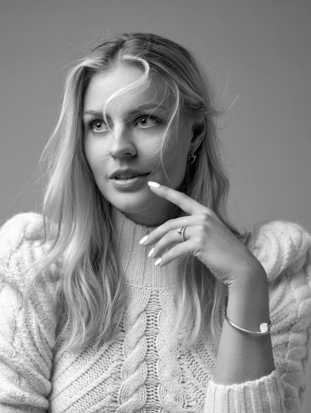 Black and white portrait of a woman with long wavy hair, wearing a knitted sweater, touching her chin with her right hand, accessorized with a bracelet, ring, and small hoop earrings.