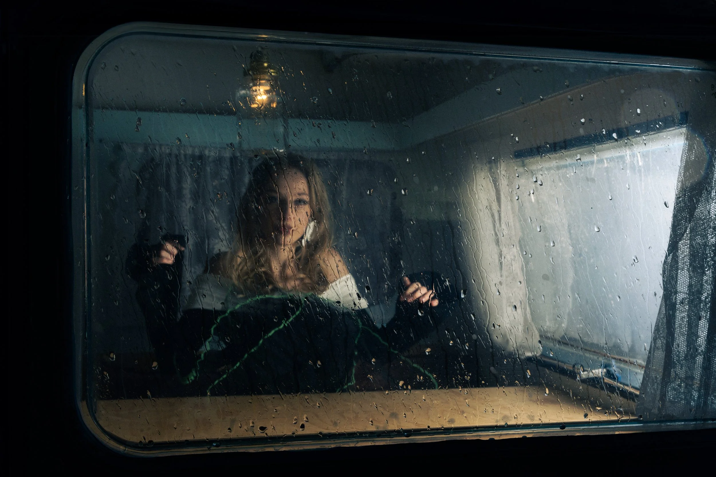 Woman inside a vehicle looking out through a rain-covered window, with a cloudy sky and a house visible outside.