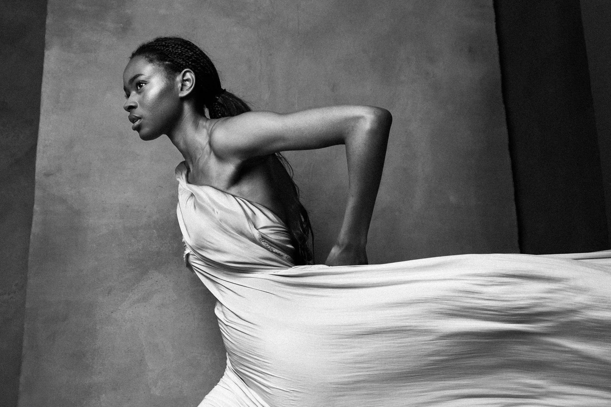 Black and white photo of a woman with braided hair, mid-movement, wearing a flowing dress, against a plain background.