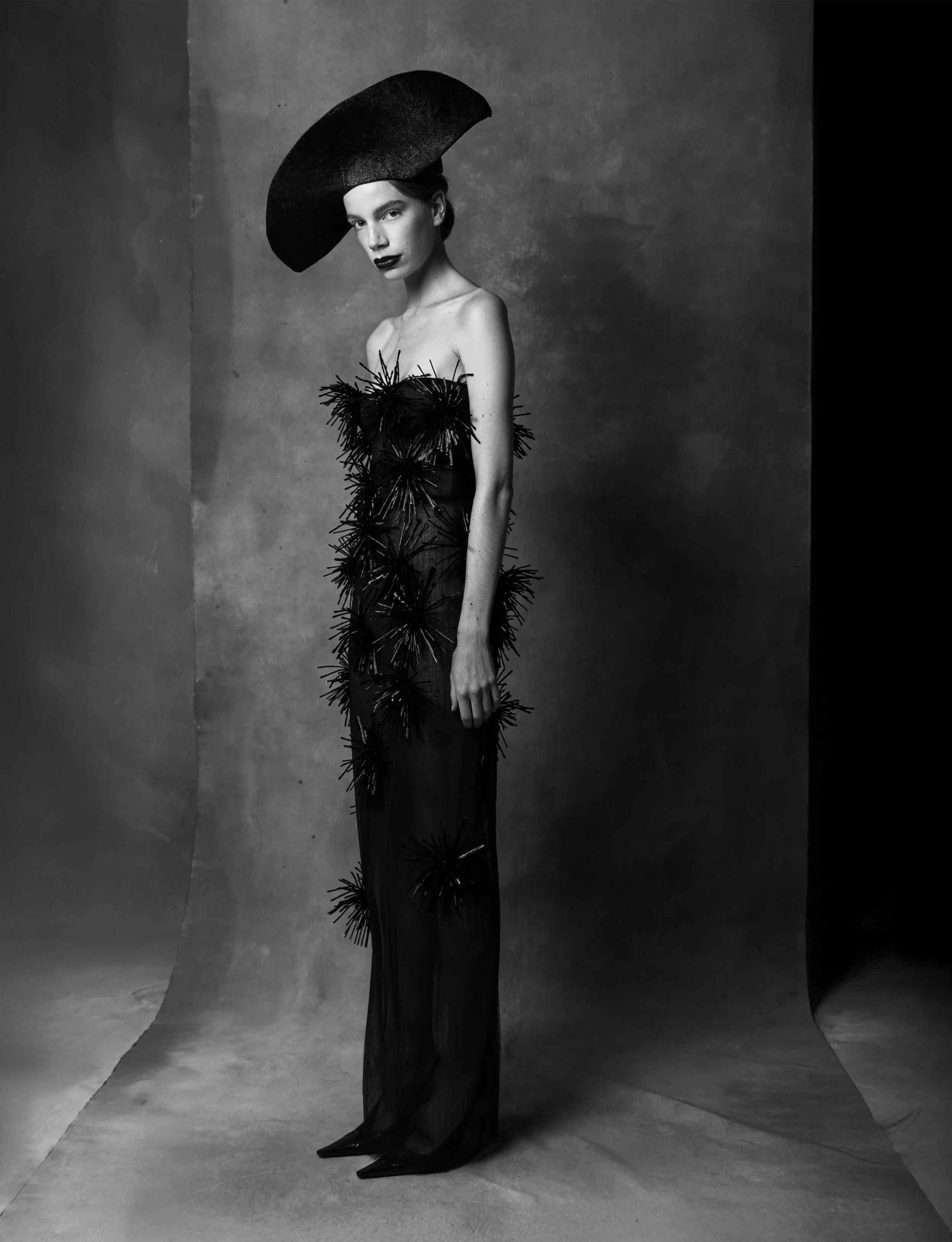 A woman in a black, strapless dress with textured, starburst-like embellishments stands against a plain backdrop. She wears a large, wide-brimmed hat and has dark lipstick. For Numero Magazine Netherlands.