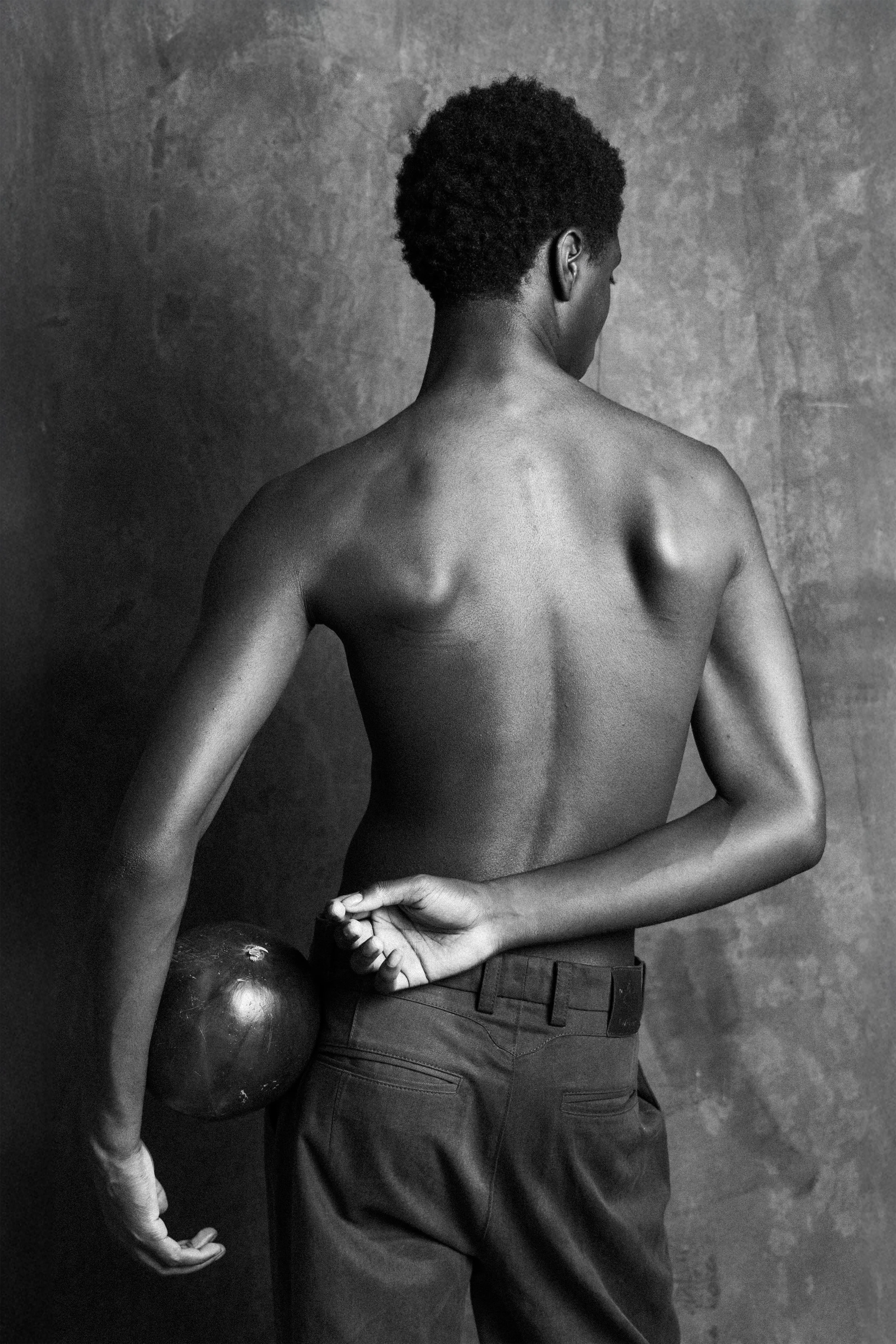 A shirtless person standing with their back to the camera, holding a medicine ball behind their back, in front of a textured wall.