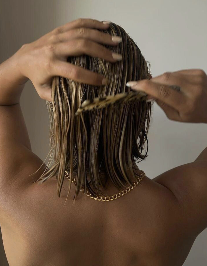 Person with shoulder-length wet hair brushing it with a wooden comb, wearing a gold chain necklace.
