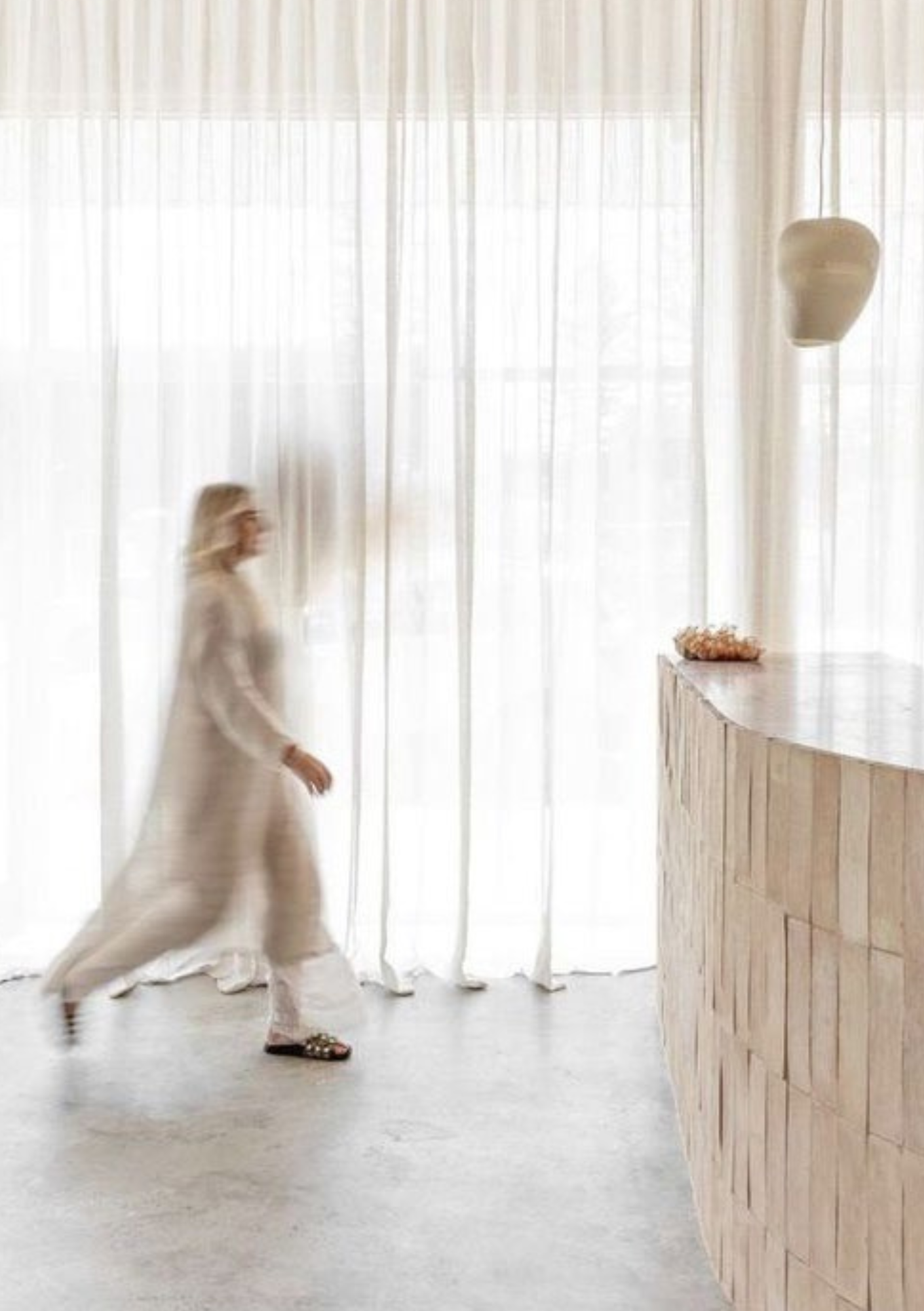 A blurred woman walking past a curved wooden reception desk with a decorative object on top, in front of a sheer curtain backdrop with diffused natural light.