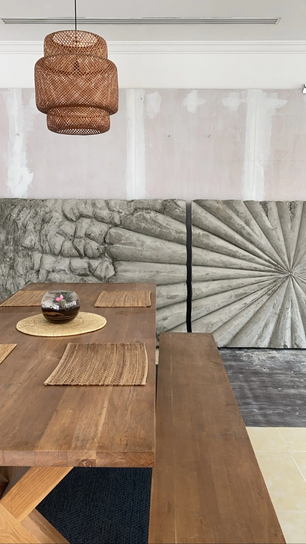 A dining room with a wooden table, four woven placemats, and a small glass bowl with plants. A large hanging woven light fixture is above, and the wall has a large decorative stone and concrete design with a swirl pattern.