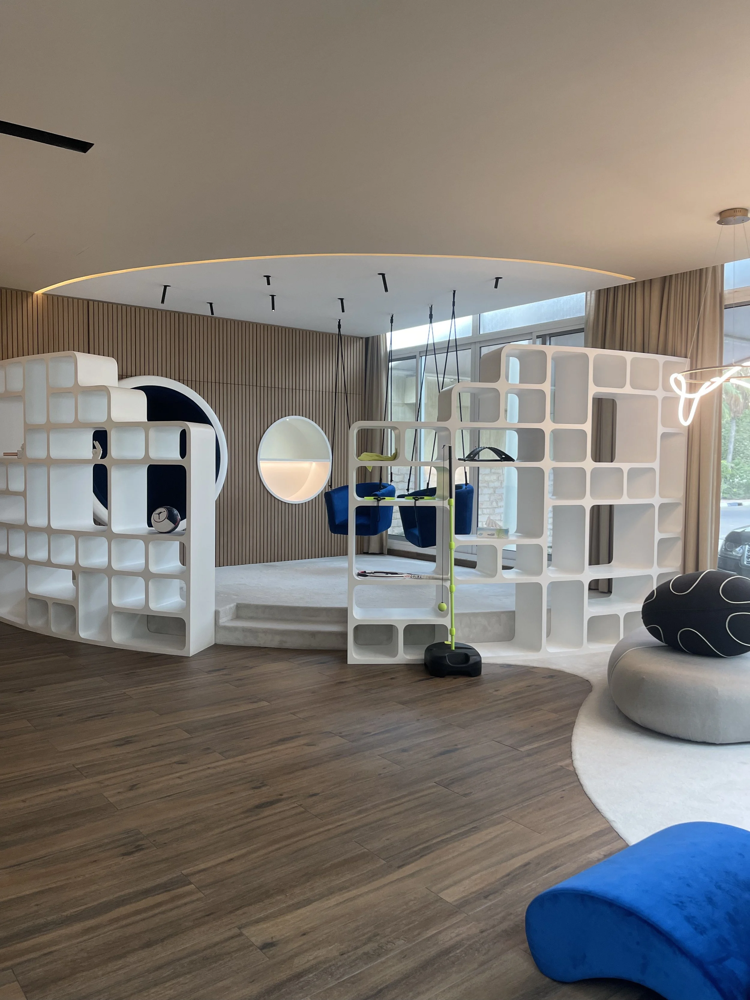 Modern indoor play area with white geometric dividers, a small stage, swings, and cozy seating with cushions and a round rug, near large windows.