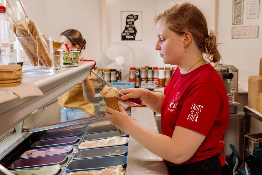 Een vrouw die ijs in een hoorntje schept in een ijssalon, met andere mensen en ingrediënten op de achtergrond.