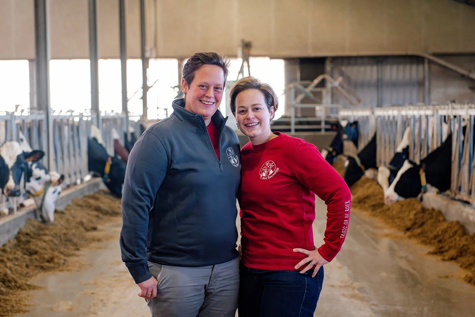 Twee mensen glimlachen in een koeienstal, omringd door kalfjes en koeien.