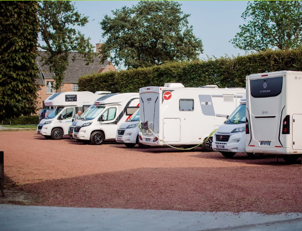 Parkeerplaats met meerdere kampeerauto's en campers op een gravel ondergrond, omgeven door bomen en huizen in de achtergrond.
