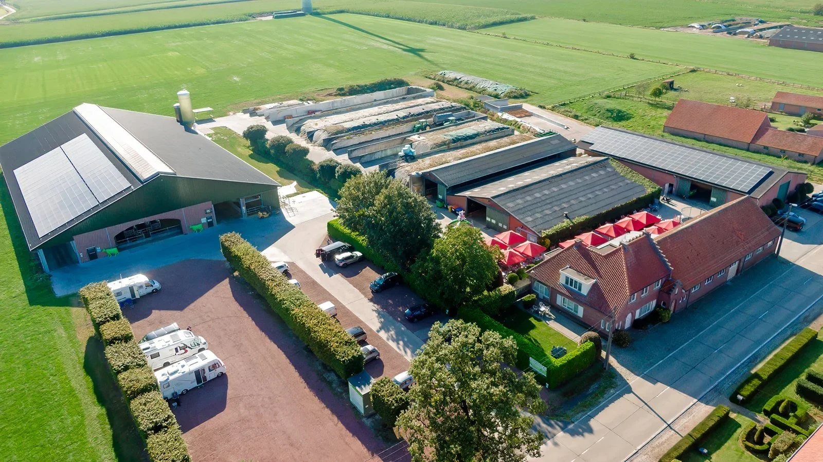 Een boerderij met verschillende schuren en panden, omgeven door groene velden en bomen, met auto's en campers in de parkeerplaatsen.