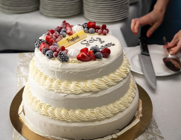 Een drie verdiepingen tellende trouwtaart met witte basis, versierd met rozenbottels, bessen en fruit, met een bericht op de bovenkant en een paar handen die een mes vasthouden klaar om te snijden.