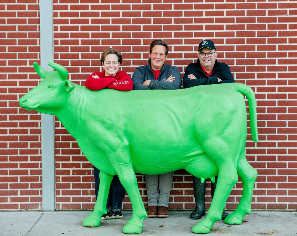 Drie mensen staan achter een grote, groene koe-vast gebonden standbeeld tegen een bakstenen muur.