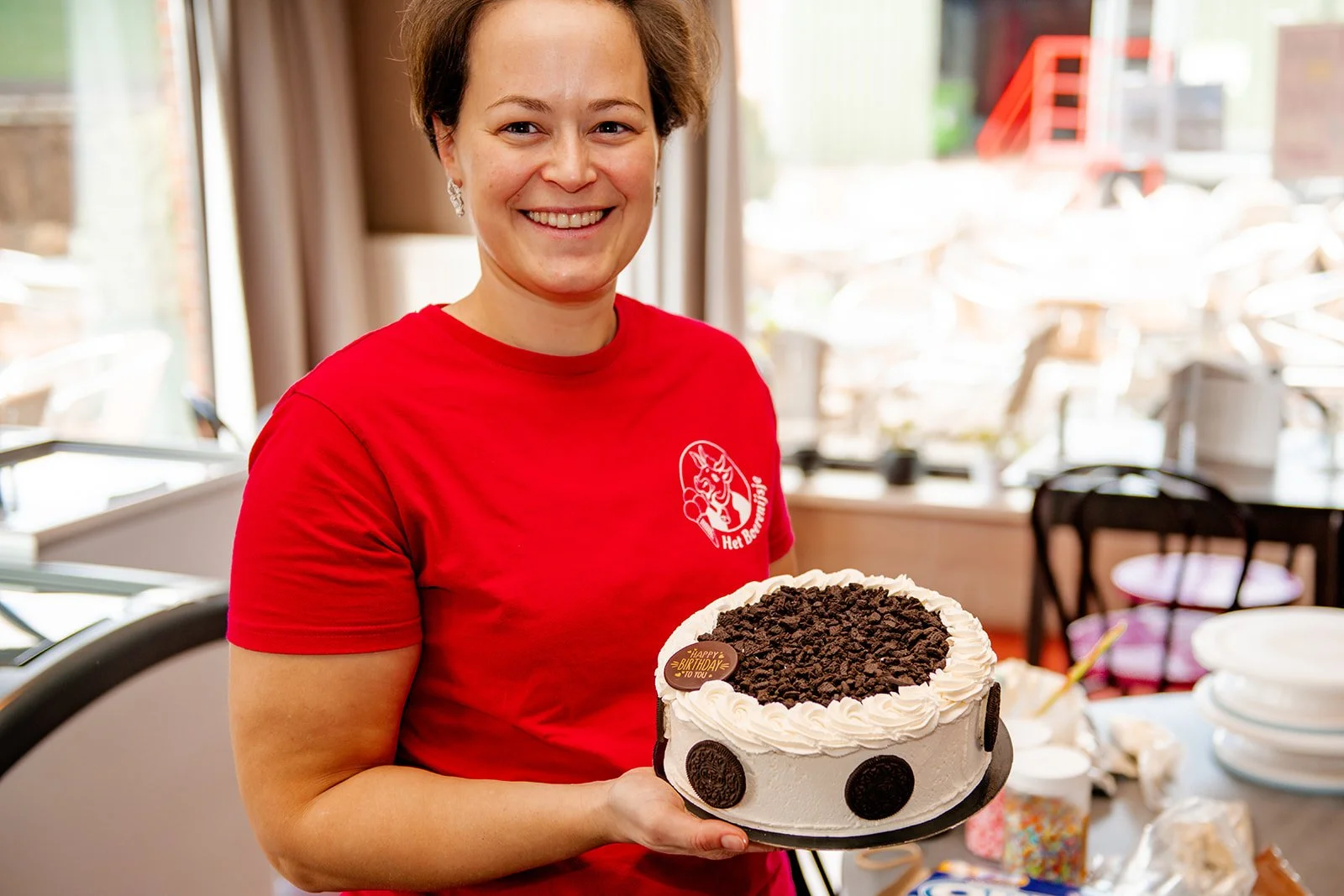 Vrouw met kort bruin haar in een rode T-shirt die een Oreo-taart vasthoudt, lachend.