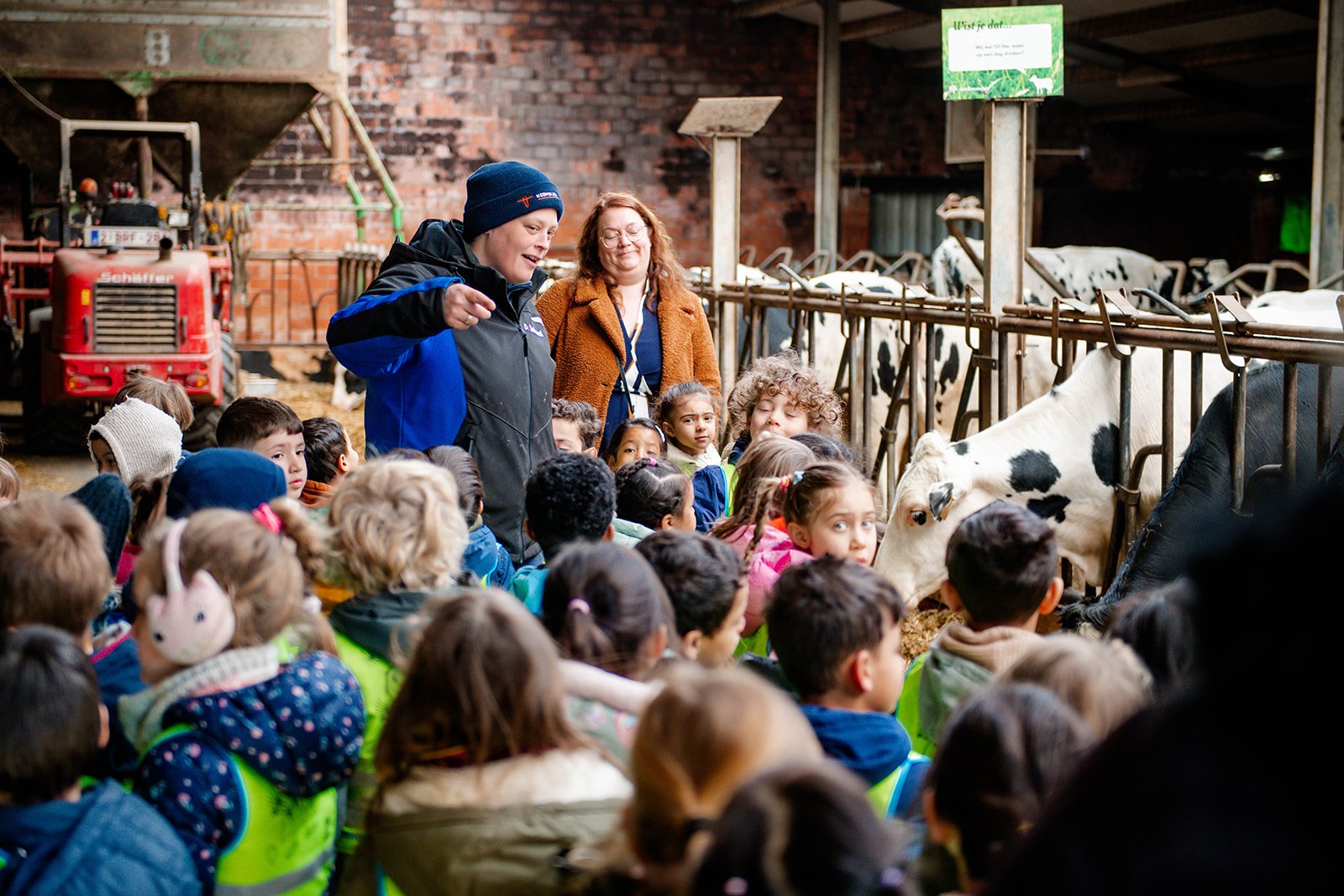 Kinderen krijgen een educatieve rondleiding over een melkveebedrijf, luisteren naar uitleg over koeien, met twee volwassen begeleiders, in een schuur met koeien en landbouwmachines op de achtergrond.