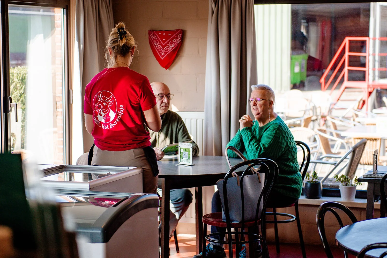 Een serveerster in een rood shirt met een logo van Het Boerenijsje spreekt met een oudere man en vrouw in een café. De man houdt een menukaart vast, terwijl de vrouw met een groene trui naar de serveerster kijkt. Grote ramen laten veel licht binnen.