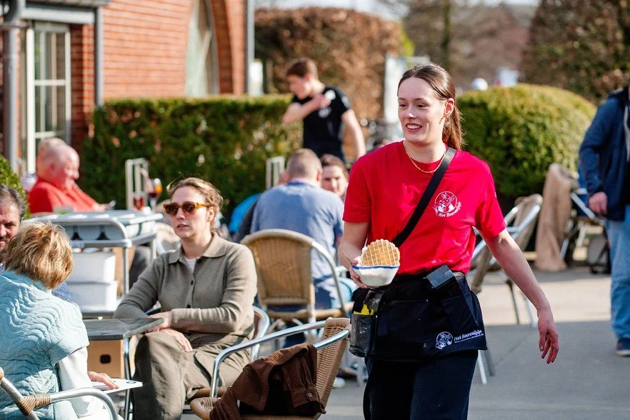 Vrouw met rode T-shirt en zwarte schort serveert een wafel in een café buiten, omringd door andere mensen die zitten en praten.