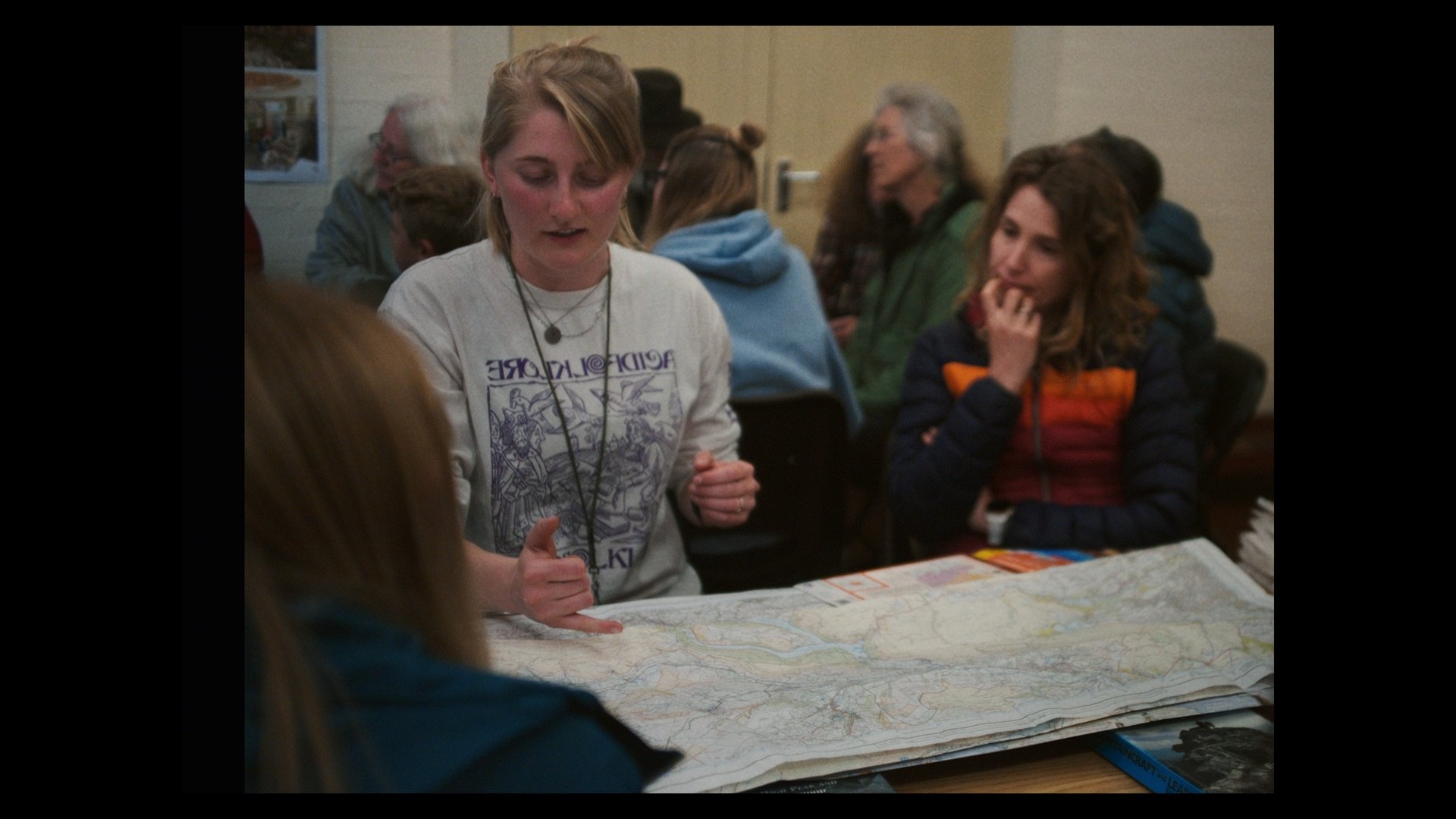 A group of people gathered around a table, with one young woman in the center pointing at a large map. Others are watching her, some with thoughtful expressions.