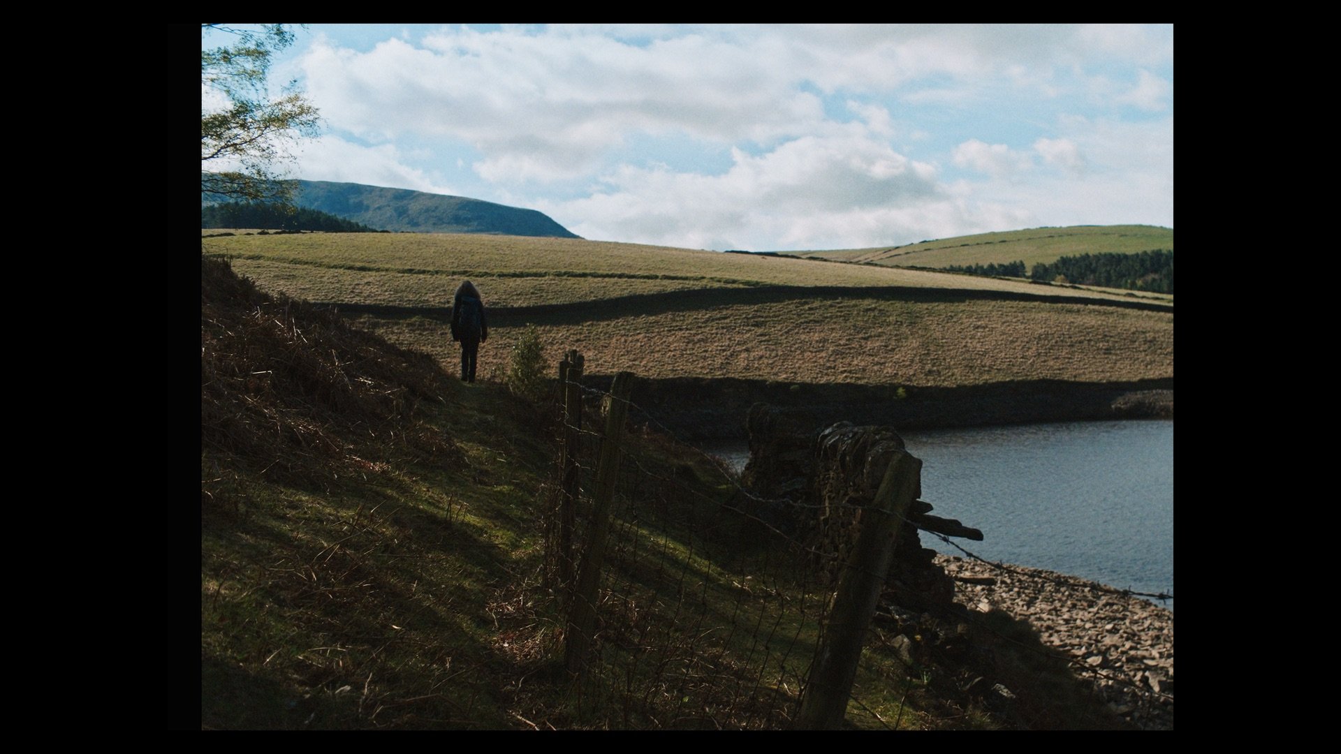 A person with a backpack walking along a fenced trail next to a body of water, with rolling grassy hills and a partly cloudy sky in the background.
