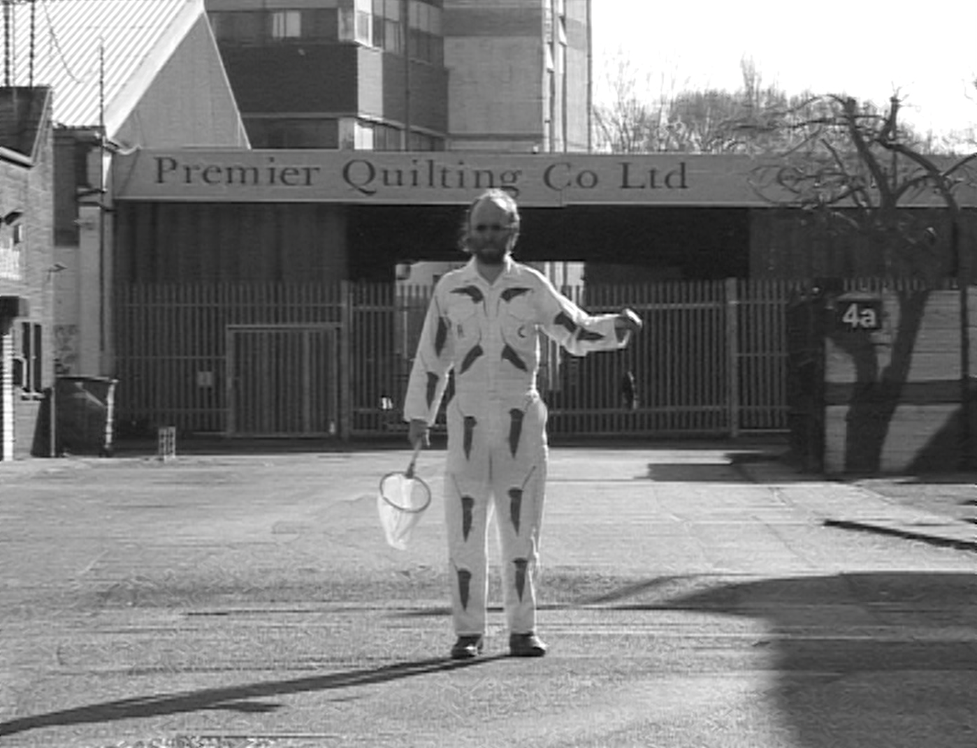 A person dressed in a cow-patterned jumpsuit holding a net and a small bag, standing on a street in front of a closed gate with a sign that reads "Premier Quilting Co Ltd".
