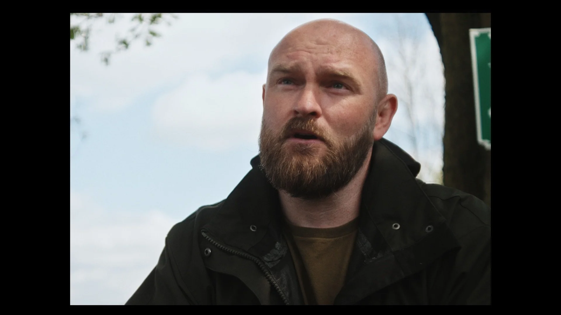 A man with a beard and shaved head, wearing a black jacket, outdoors during the day with a cloudy sky and trees in the background.