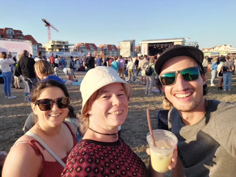 Drie mensen lachen samen op een festivalterrein in Nieuwpoort, met een podium en andere festivalgangers op de achtergrond, zichtbaar genietend van muziek en elkaars gezelschap.