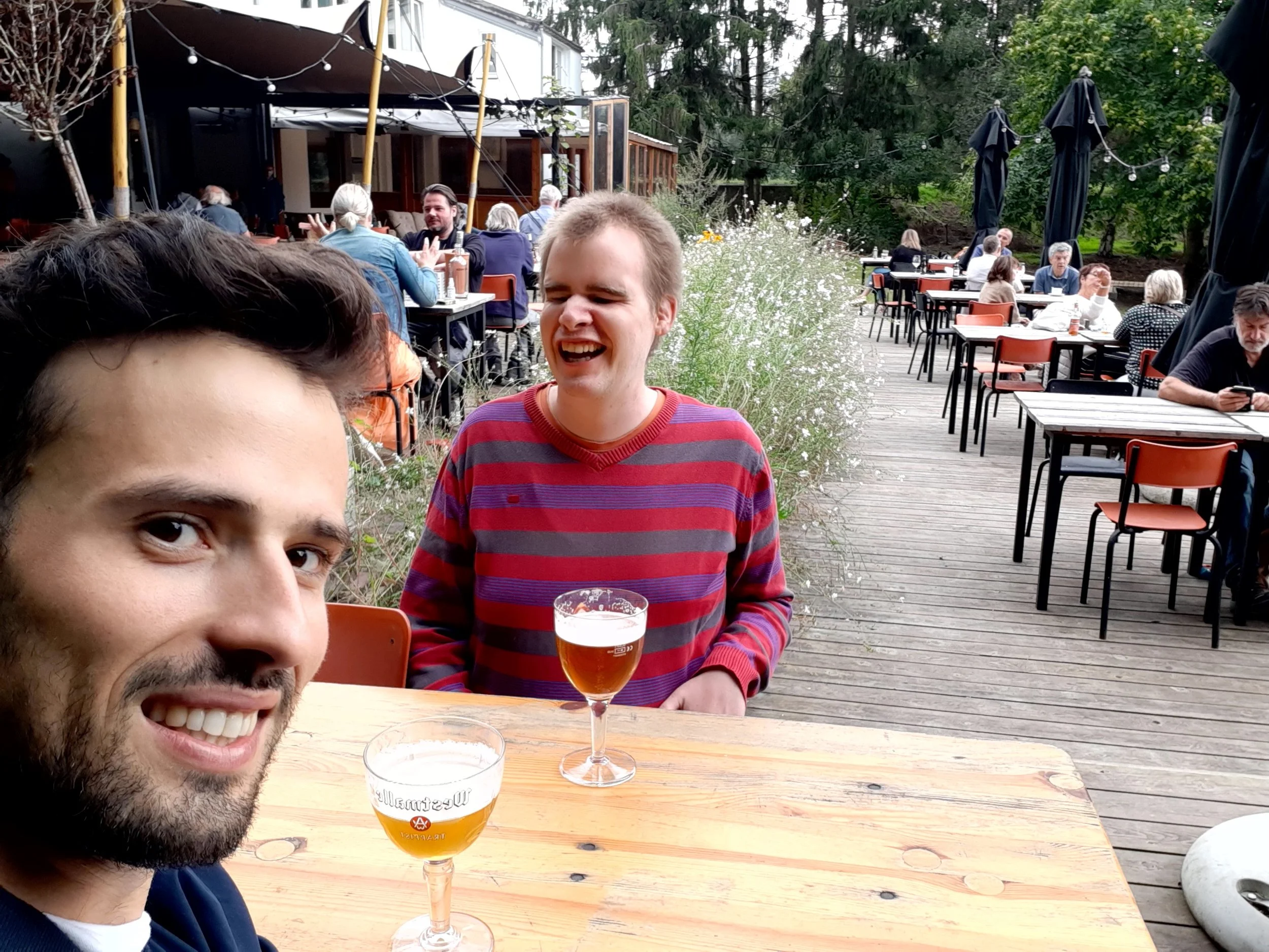 Twee volwassenen zitten samen aan een houten tafel op een gezellig terras, genieten van een drankje en lachen, met andere gasten en groen op de achtergrond.