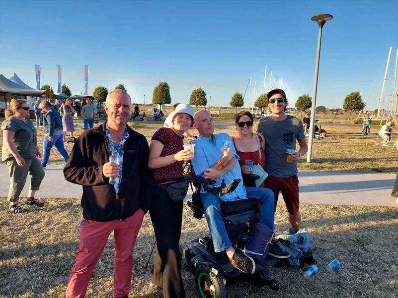 Een groep volwassenen poseert samen op een zonnig festivalterrein, met drankjes in de hand en een rolstoelgebruiker in het midden, zichtbaar genietend van het moment en elkaars gezelschap.