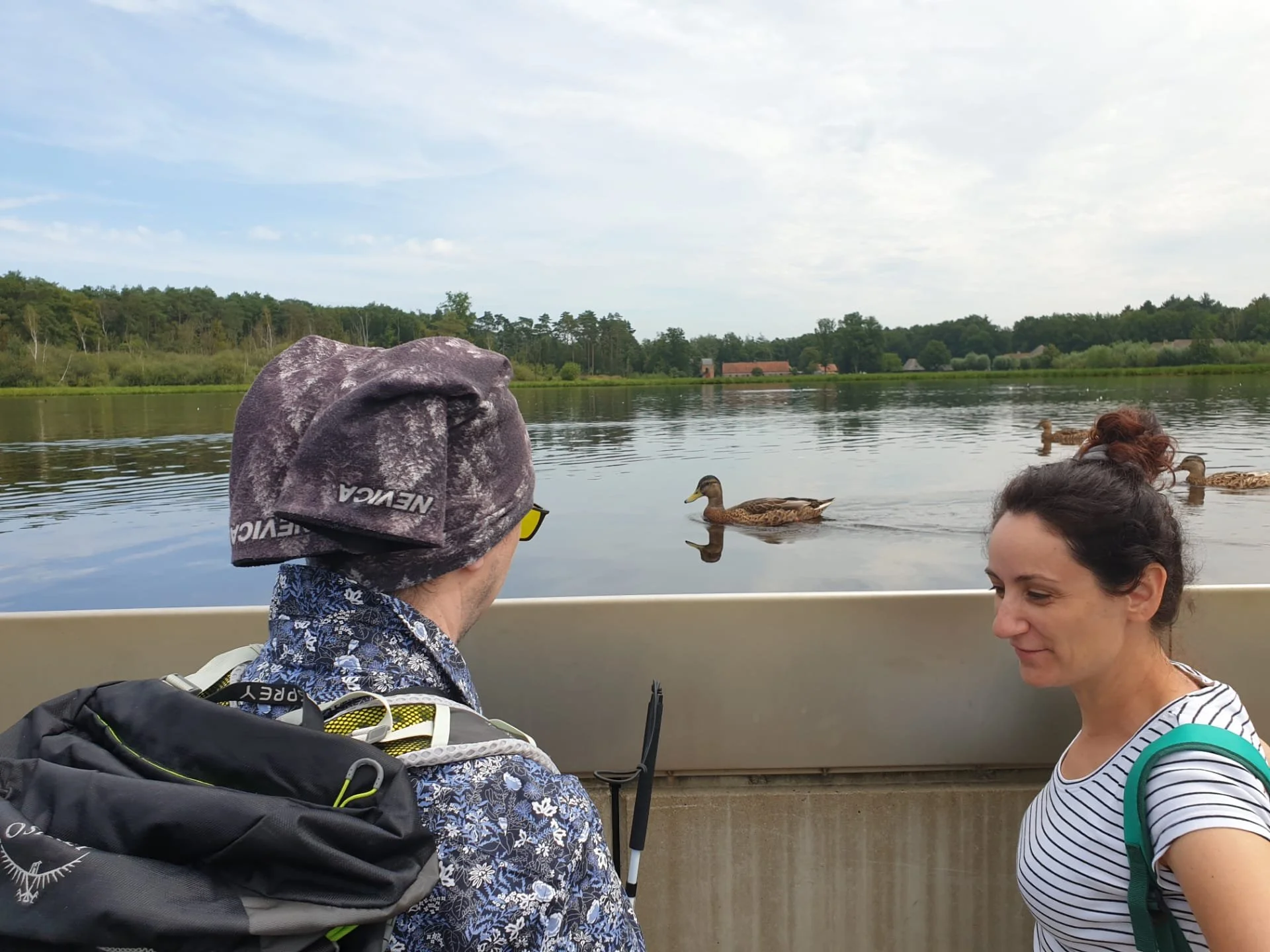 Twee personen staan samen aan een reling bij een rustig meer en kijken naar eenden op het water, met natuur en bomen op de achtergrond.