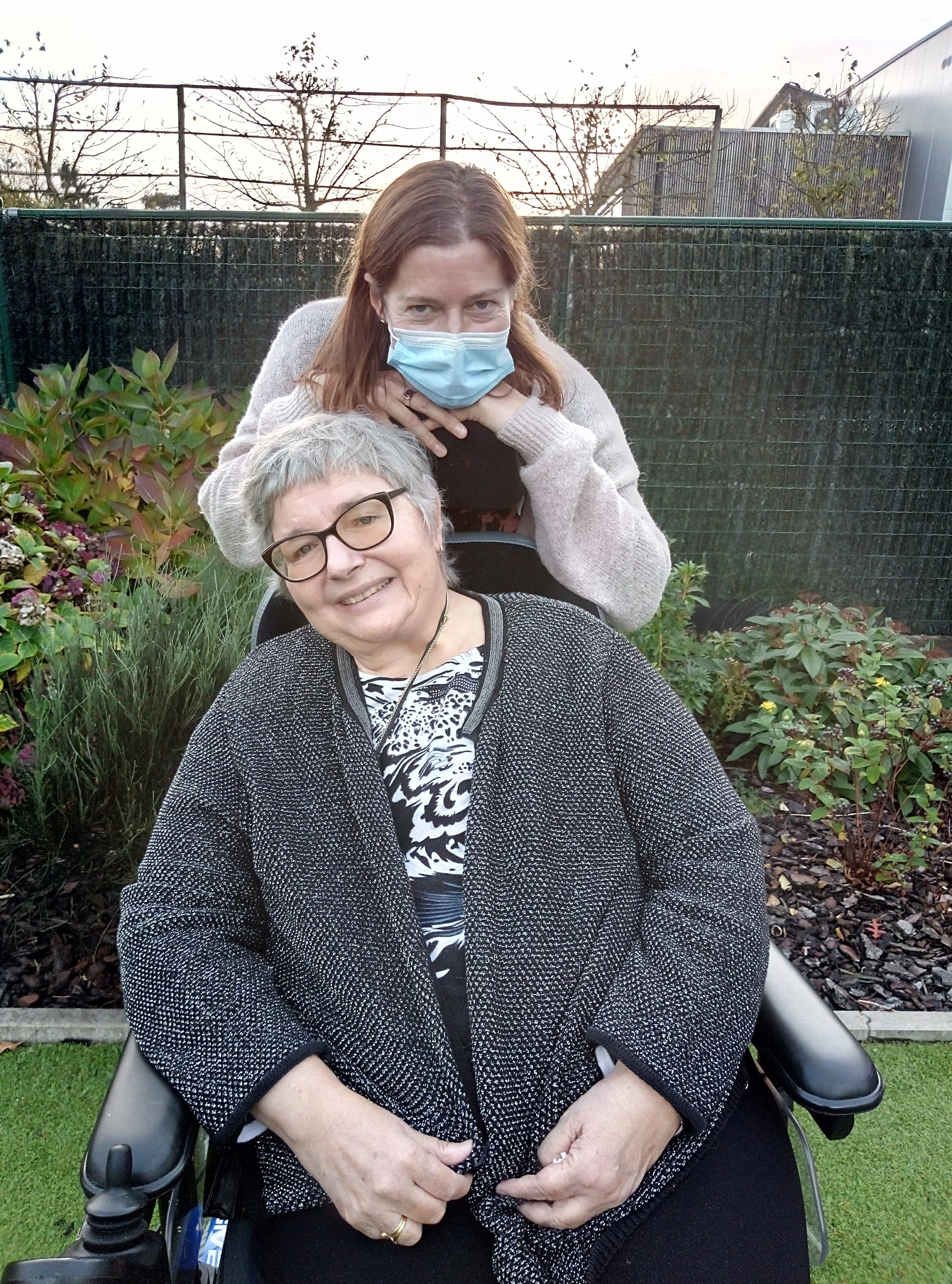 Twee vrouwen poseren samen in een tuin. Eén vrouw zit in een rolstoel en glimlacht naar de camera, terwijl de andere vrouw achter haar staat met haar handen op de rugleuning, in een warme en vertrouwde houding.
