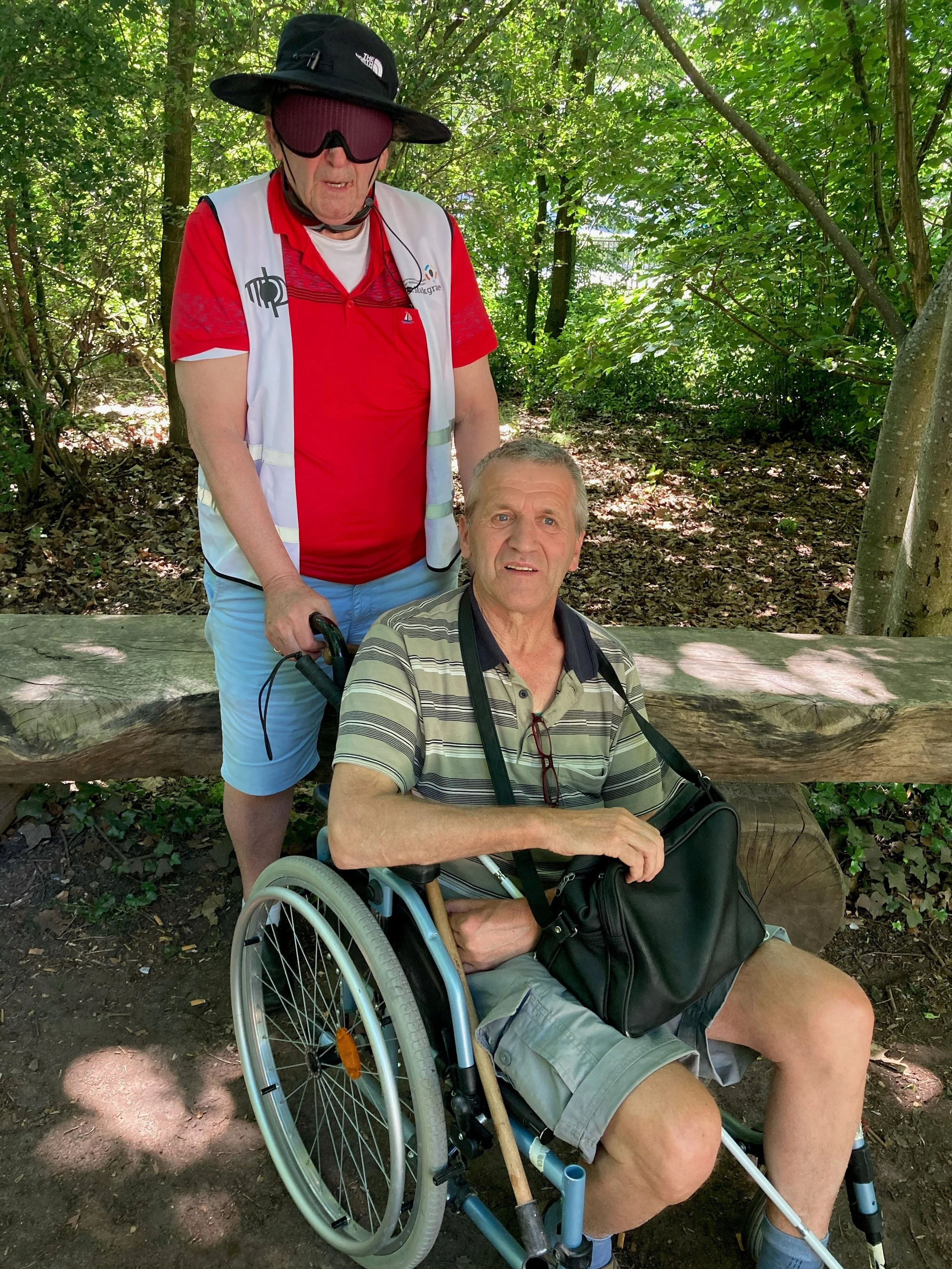 Twee mannen zijn samen in een bosrijke omgeving. Eén man zit in een rolstoel op een bospad, terwijl de andere man achter hem staat en hem begeleidt, met bomen en groen rondom hen.