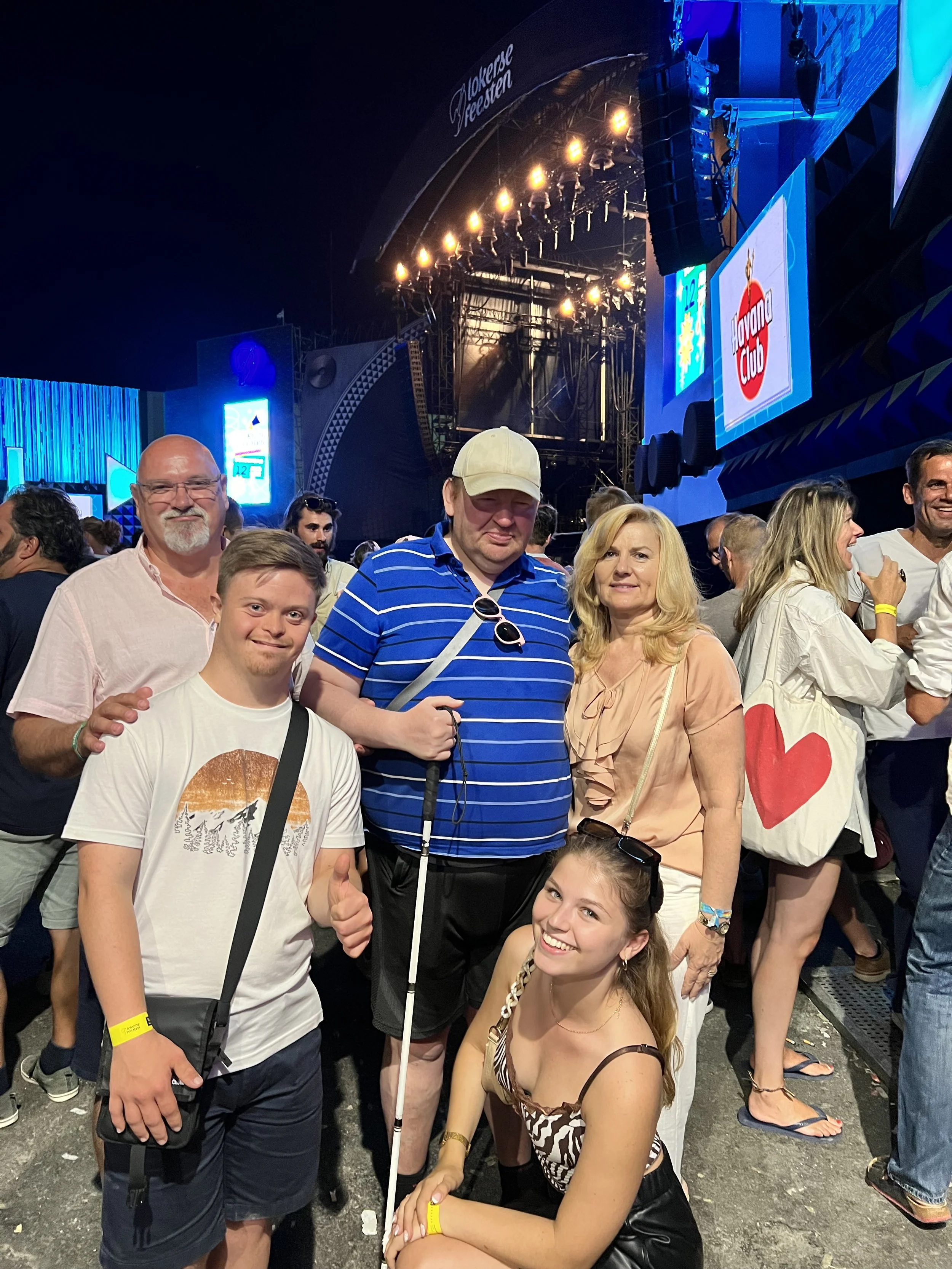 Een groep volwassenen poseert samen op een drukbezocht festivalterrein voor een verlichte podiumachtergrond, zichtbaar ontspannen en verbonden tijdens een gezamenlijke avond uit.