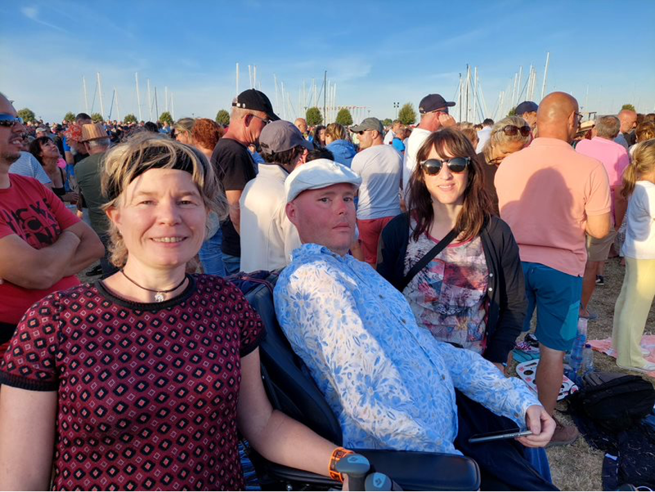 Een groep volwassenen geniet samen van een drukbezocht festival op Nostalgie Beach, met een rolstoelgebruiker op de voorgrond en een enthousiaste menigte en zomerse sfeer op de achtergrond.