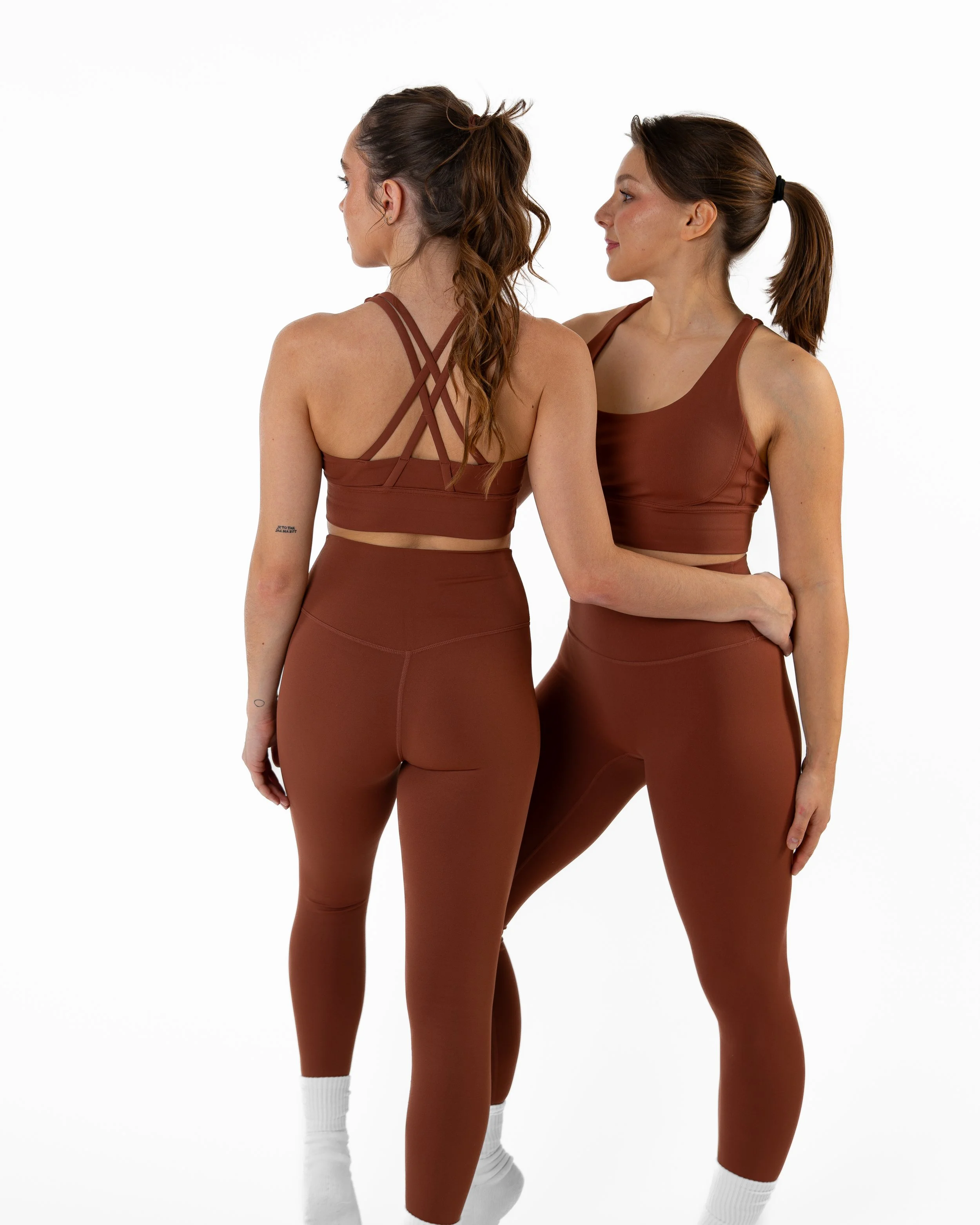 Two women wearing matching brown activewear leggings and sports bras, standing back to back against a white background.
