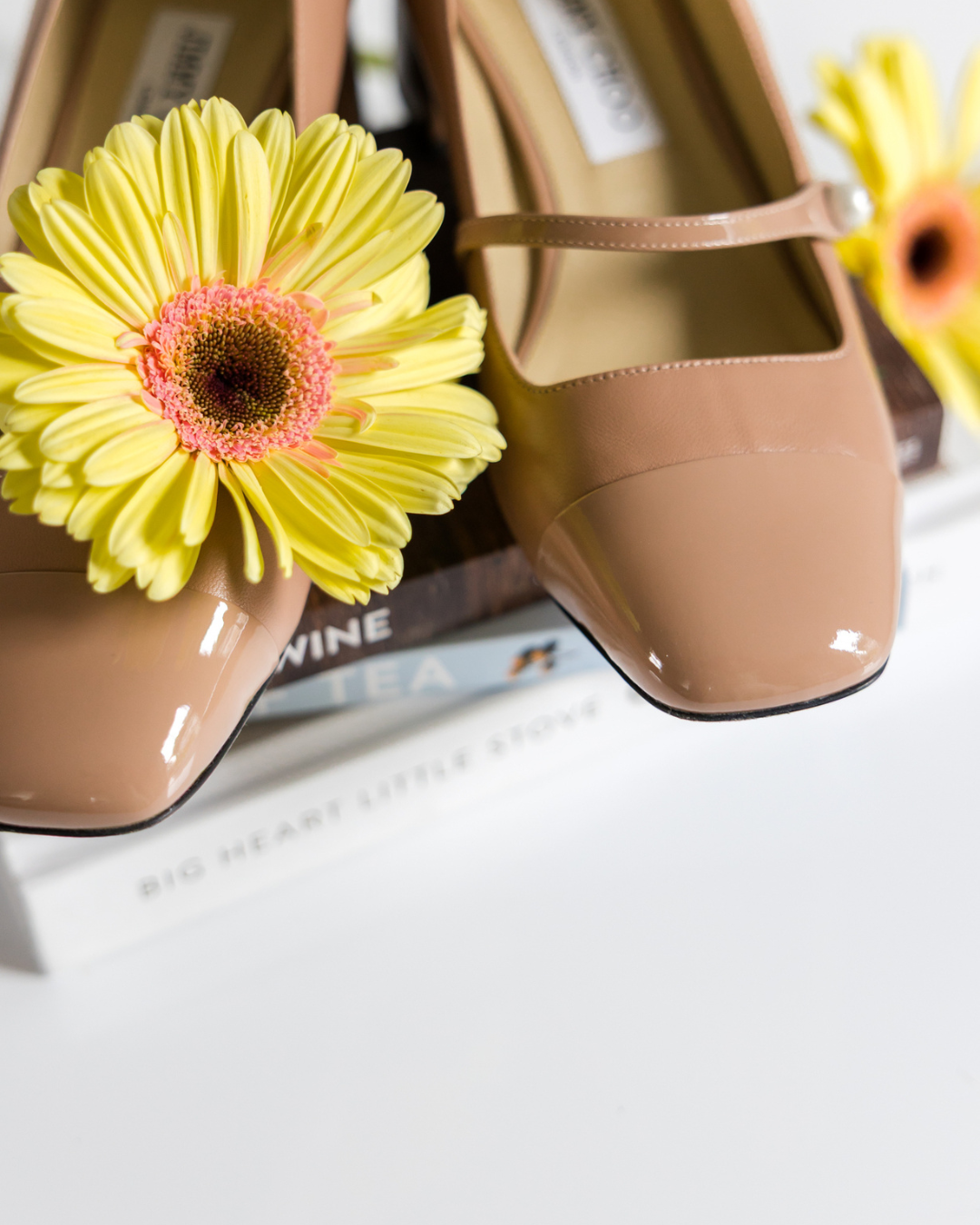 A pair of beige patent leather shoes with rounded toes, placed on a white surface next to a yellow gerbera daisy flower.